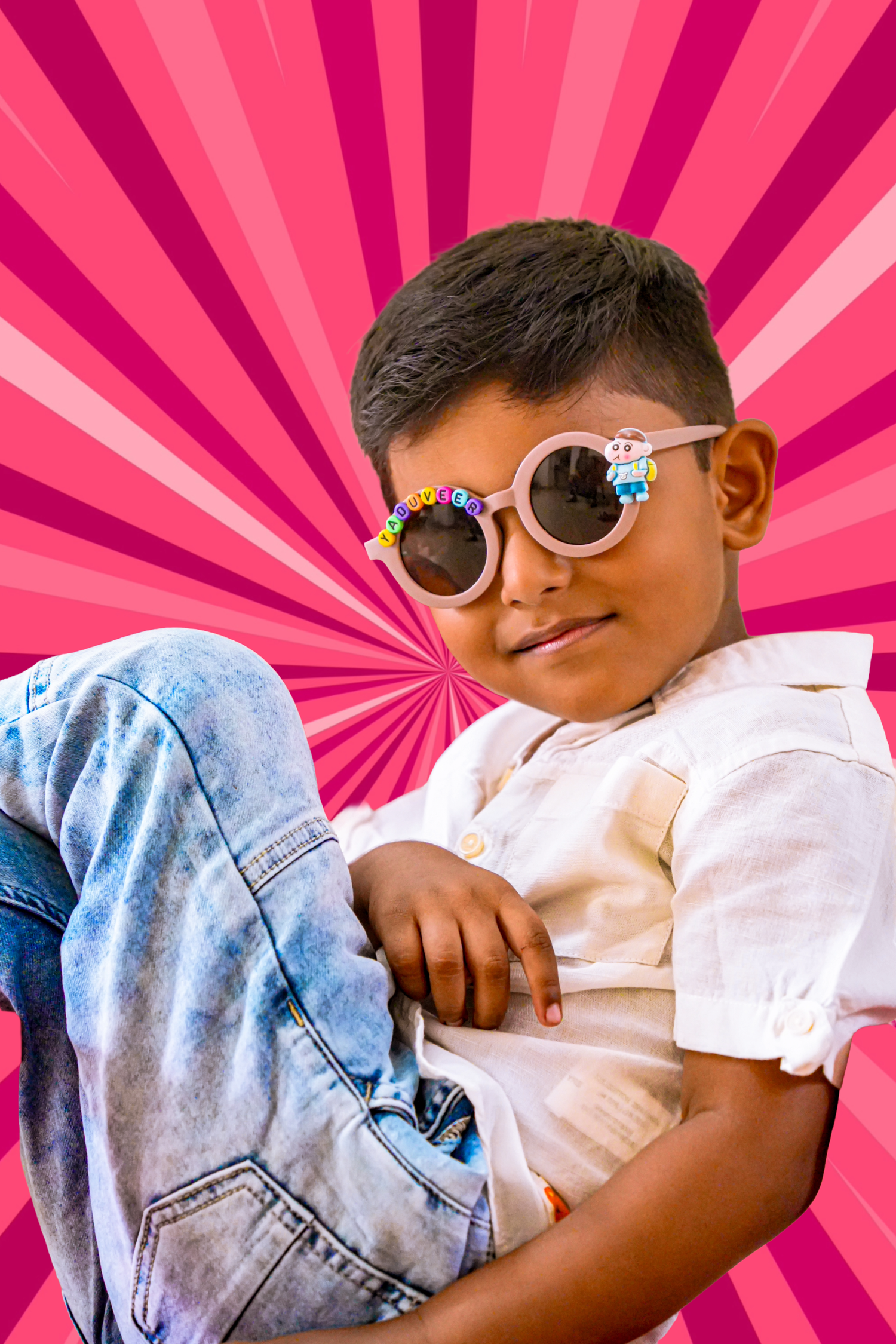 A cheerful young boy wearing decorative round sunglasses poses confidently against a vibrant pink starburst background.