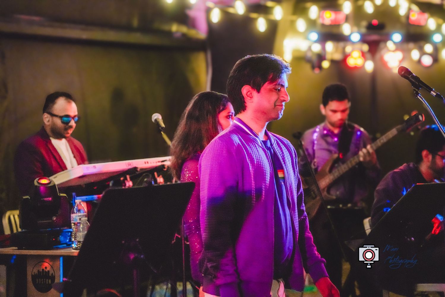 Band performing live on stage with colorful string lights, featuring a keyboardist, guitarist, and vocalist in a purple hoodie.
