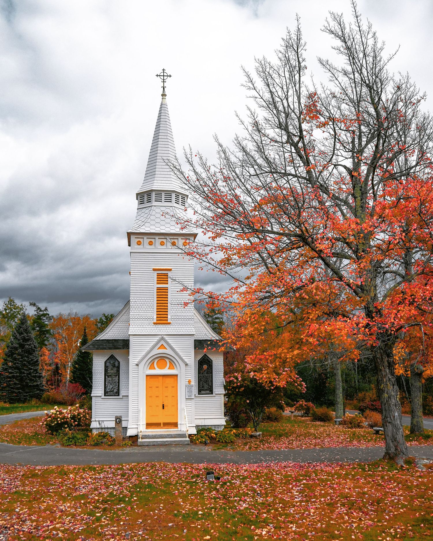 Small white church with steeple and cross surrounded by vibrant red autumn foliage under dramatic cloudy sky.