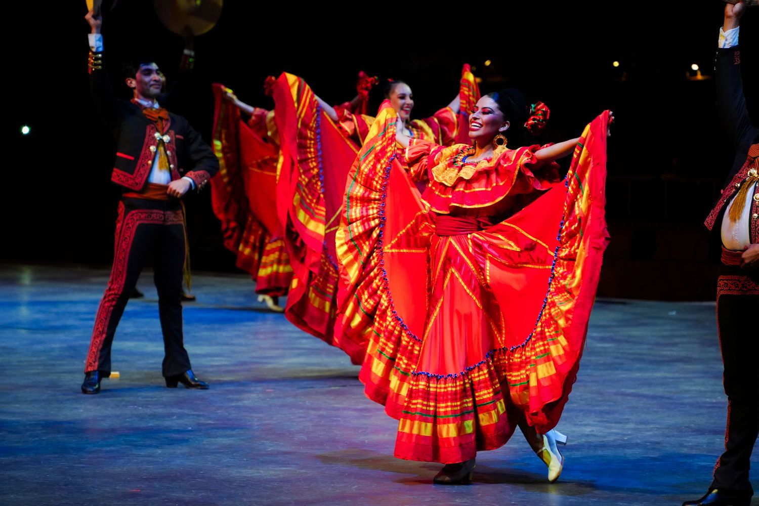 Performers in vibrant red traditional Mexican dresses dance on stage during a colorful folkloric ballet performance.