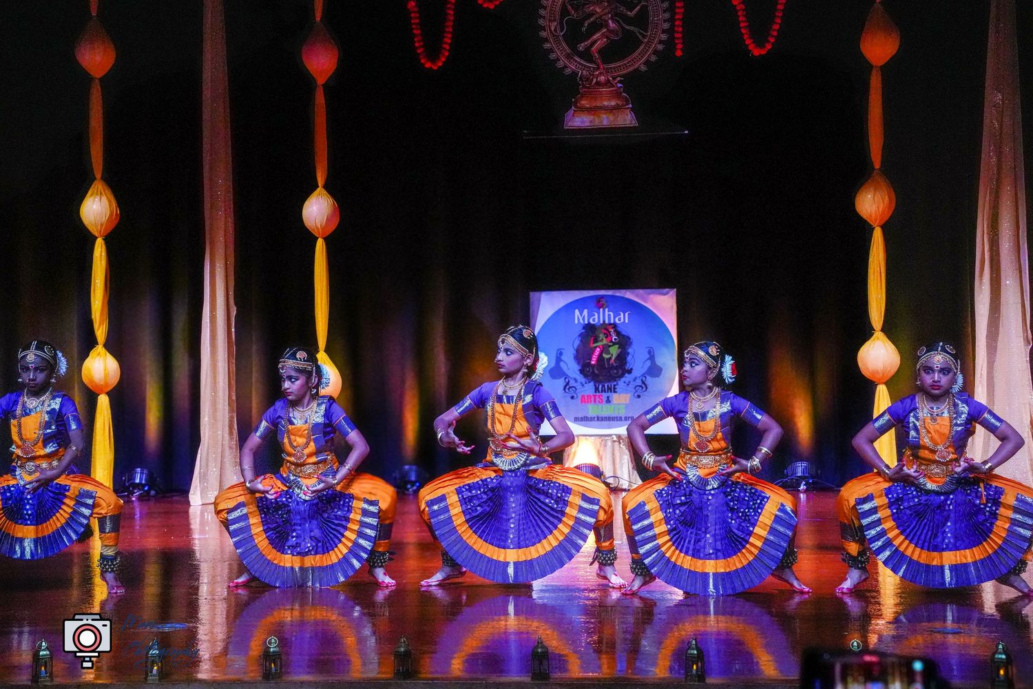 Classical Indian Bharatanatyam dancers in blue and orange costumes performing on a decorated stage.