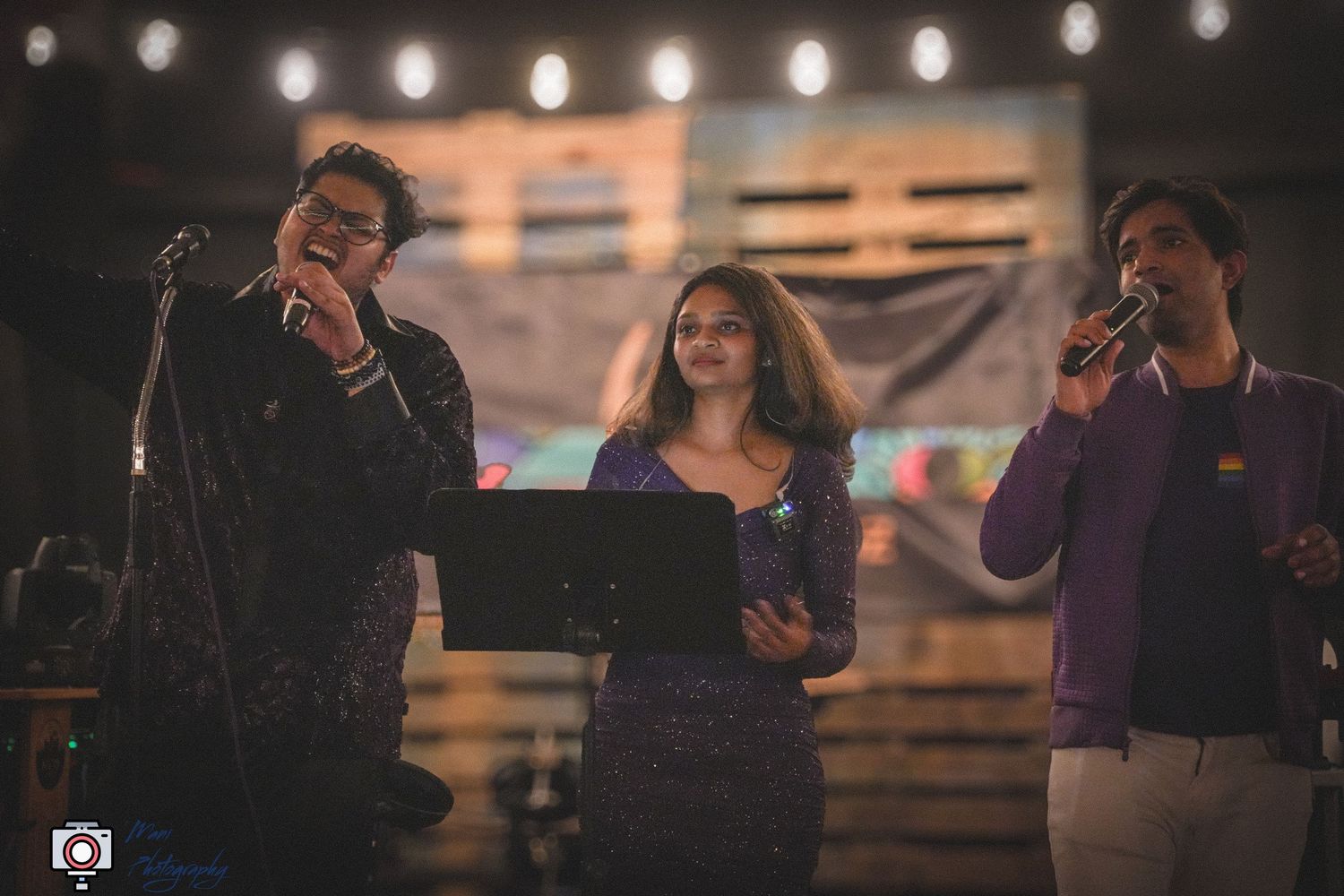 Three performers on stage with string lights at an outdoor evening event.
