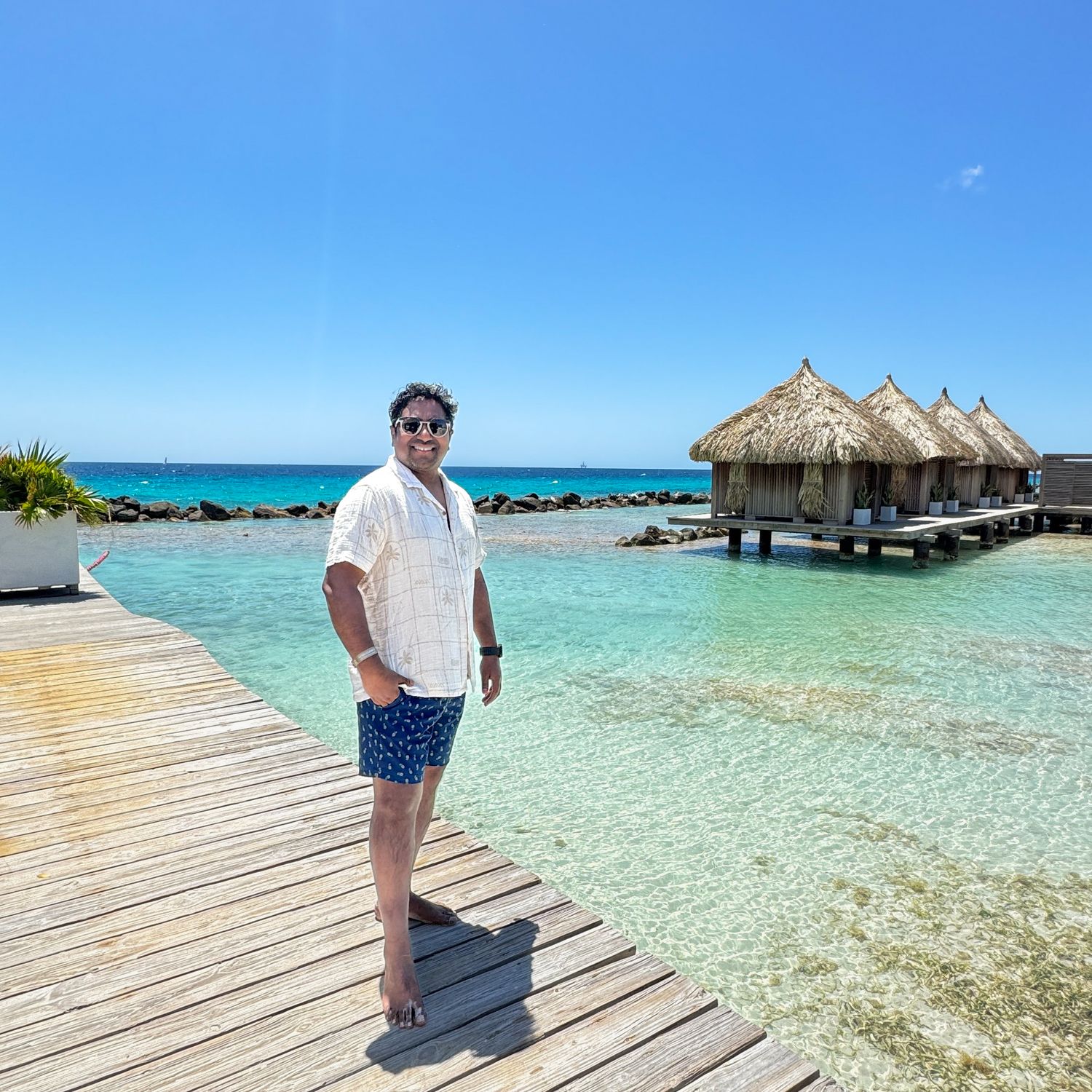 Man in white shirt stands on wooden dock with crystal clear turquoise water and overwater bungalows.