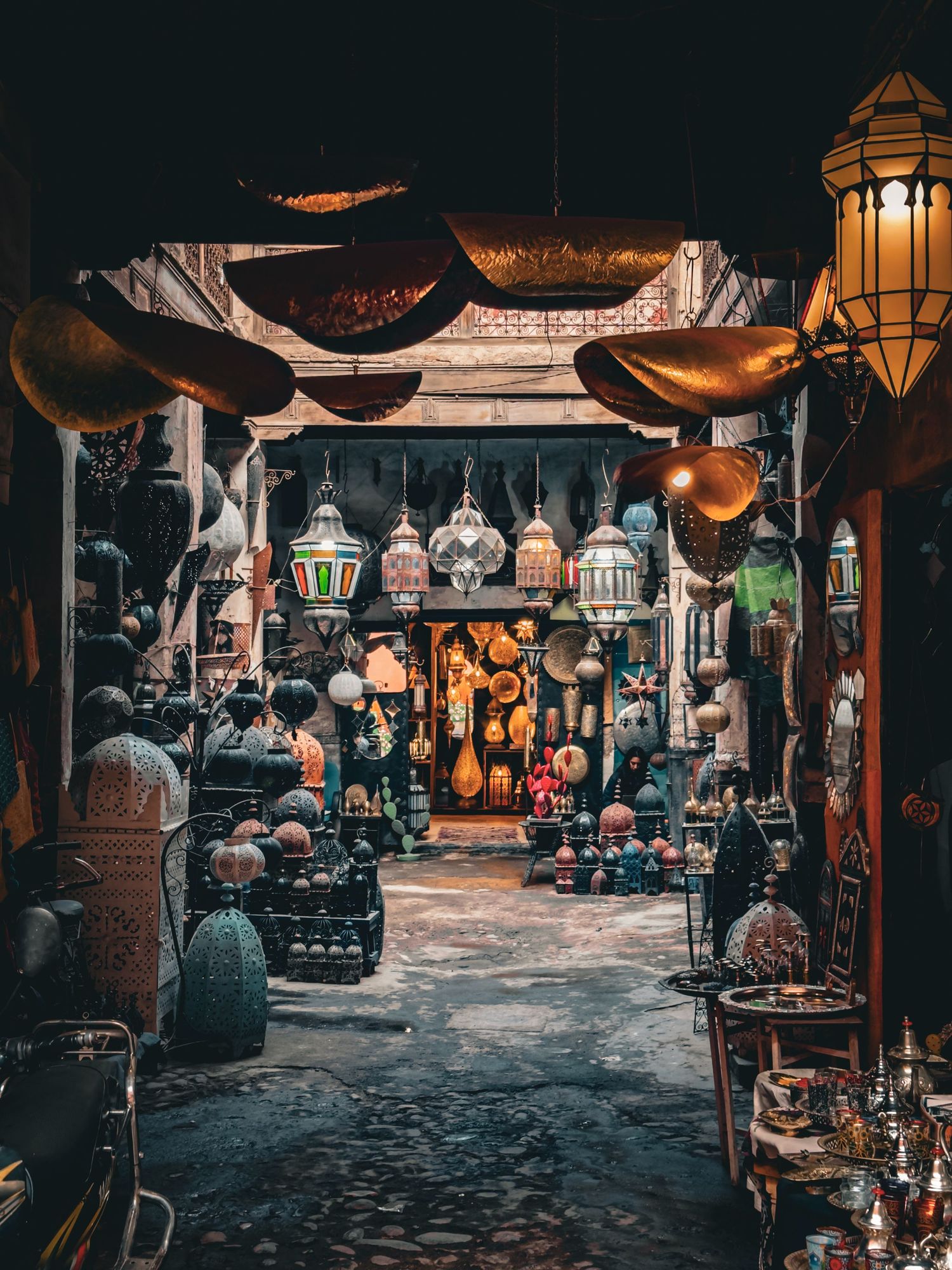 A vibrant Moroccan souk filled with ornate hanging lanterns, brass trays, and colorful pottery lining a narrow market alley.