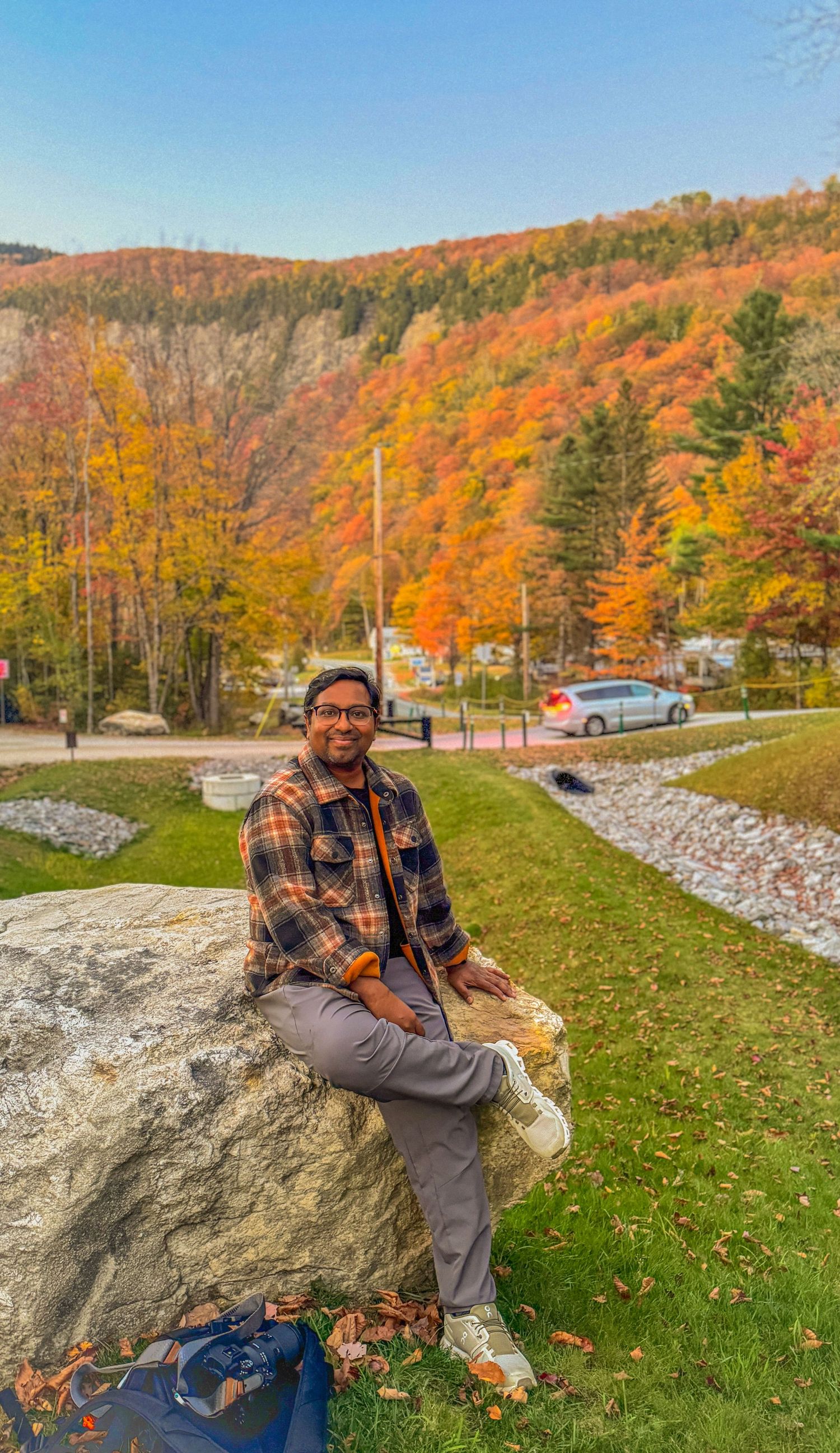 Smiling man in plaid jacket sitting on large rock with vivid autumn mountain scenery.