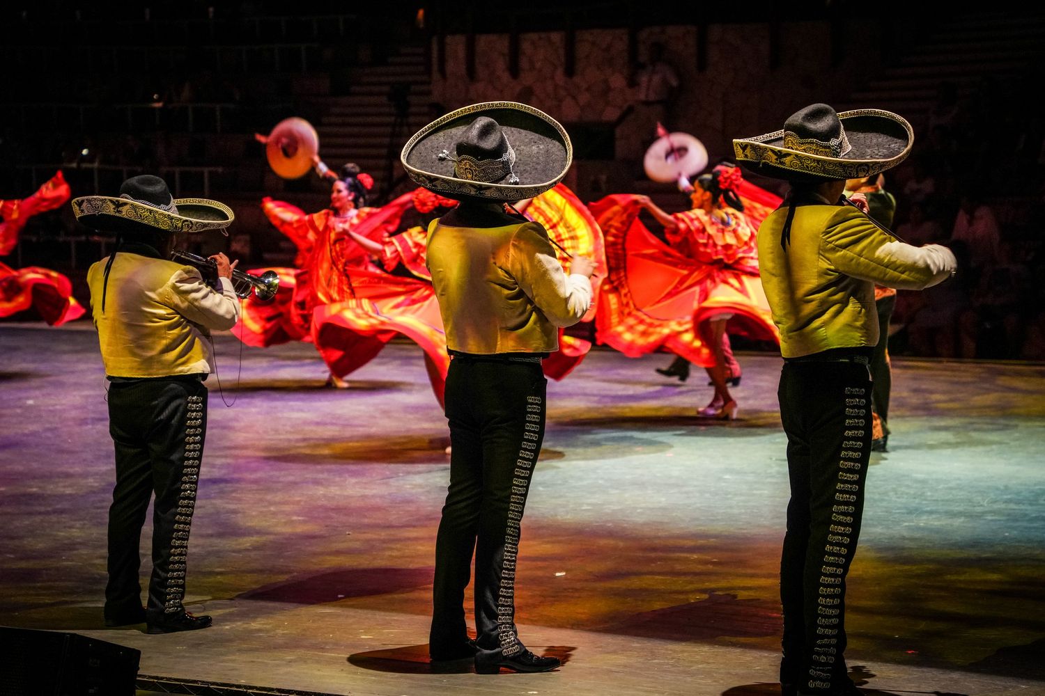 Mariachi musicians in traditional charro suits perform alongside dancers in vibrant red skirts on a colorful stage.