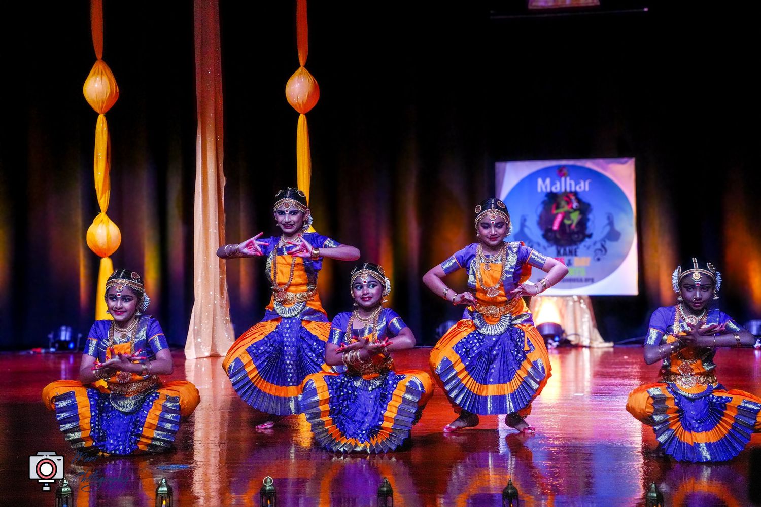 Young girls in vibrant blue and orange costumes performing Indian classical Bharatanatyam dance on a colorful stage.