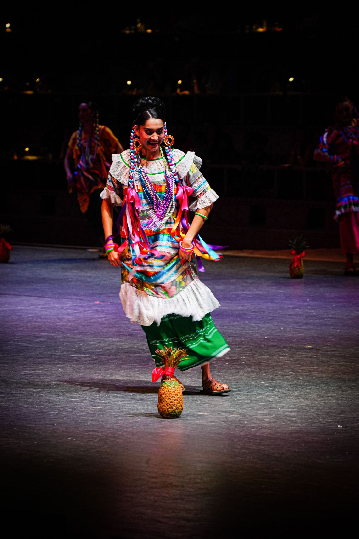 Mexican folkloric dancer in colorful traditional costume balancing a pineapple on stage during a cultural performance.