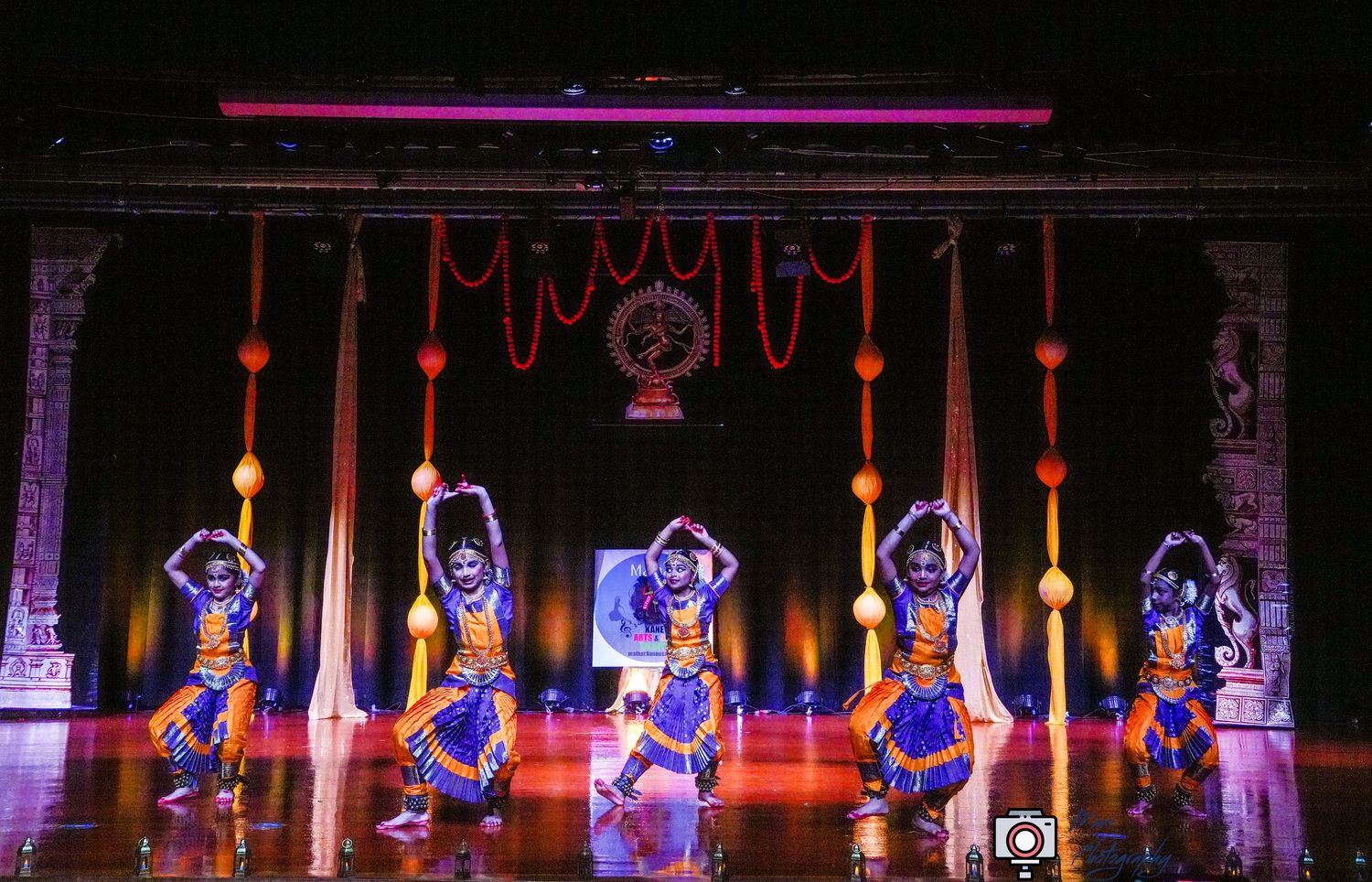 Indian classical dancers perform Odissi dance on a colorful stage with traditional temple sculptures and decorative hangings.