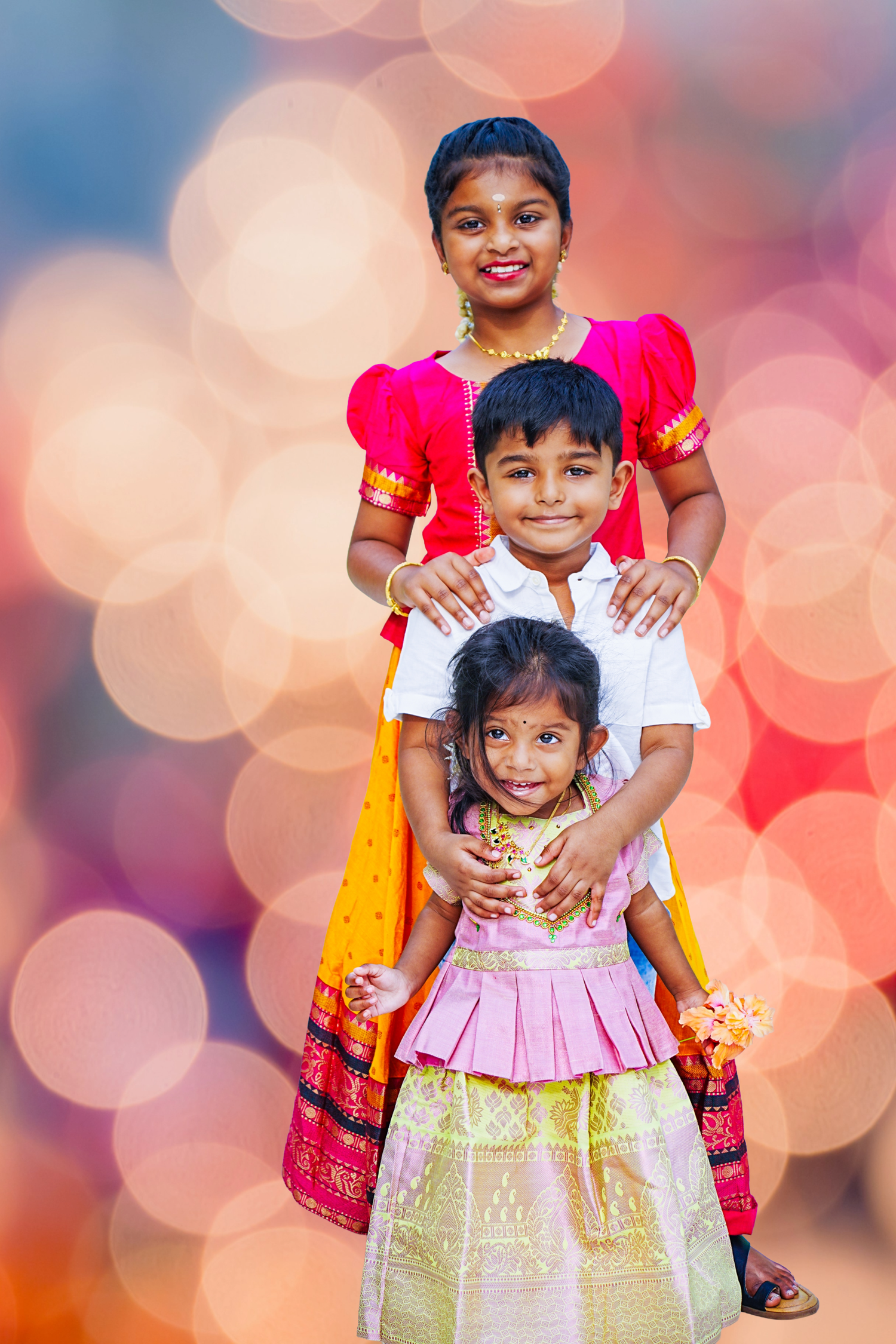 Three smiling Indian children in traditional festive attire posing together with bokeh background.