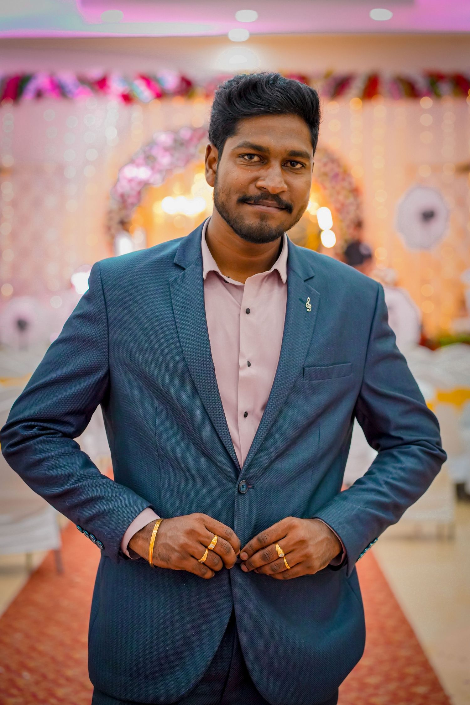 Young Indian man in blue suit adjusting jacket at decorated wedding venue.