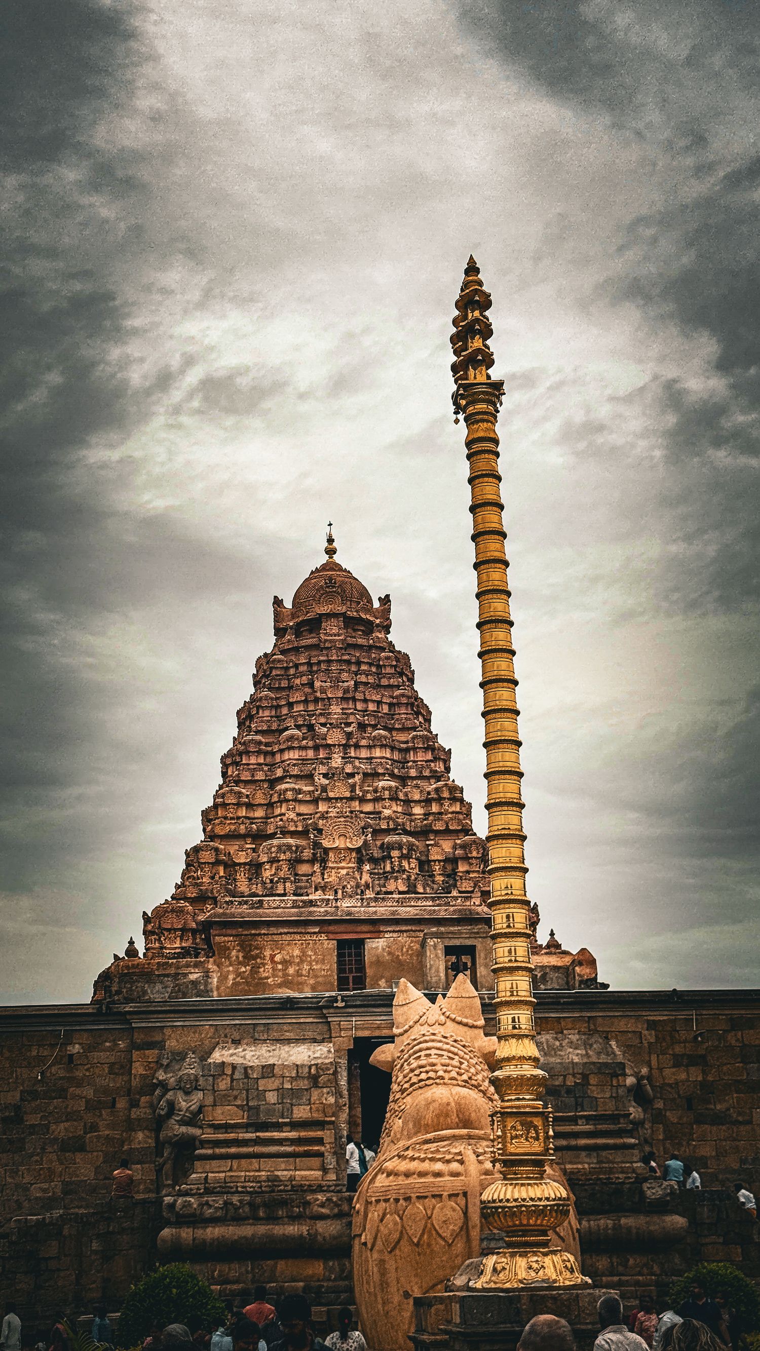 Ancient Dravidian temple gopuram with ornate stone carvings, tall golden flagpole, and Nandi sculpture under dramatic cloudy sky.