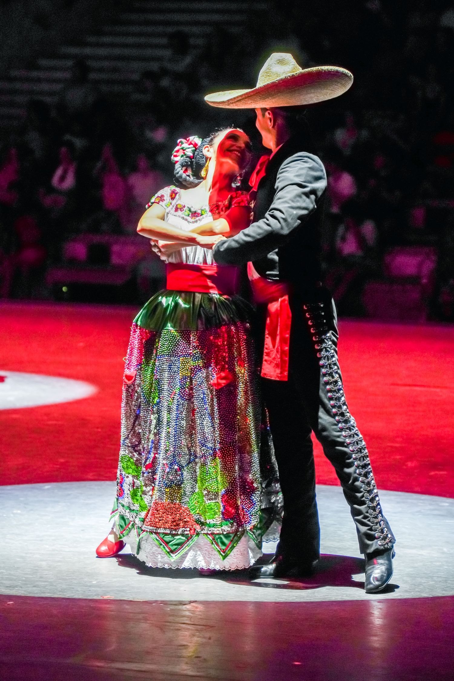 Couple in traditional Mexican folkloric costumes dancing, woman in floral dress, man in charro suit and sombrero.