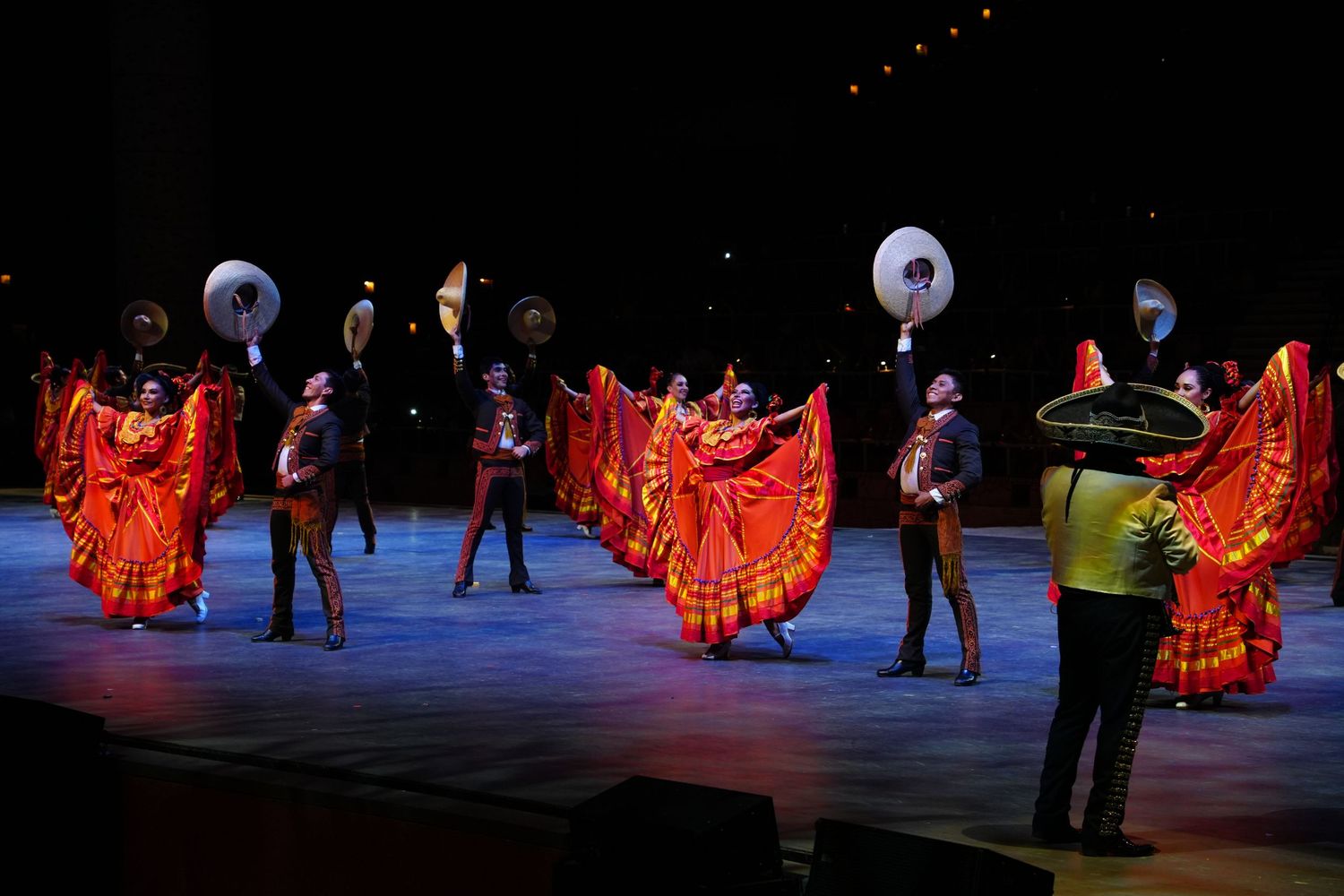 Colorful Mexican folk dancers in red dresses perform on stage with mariachi musicians in traditional charro suits.