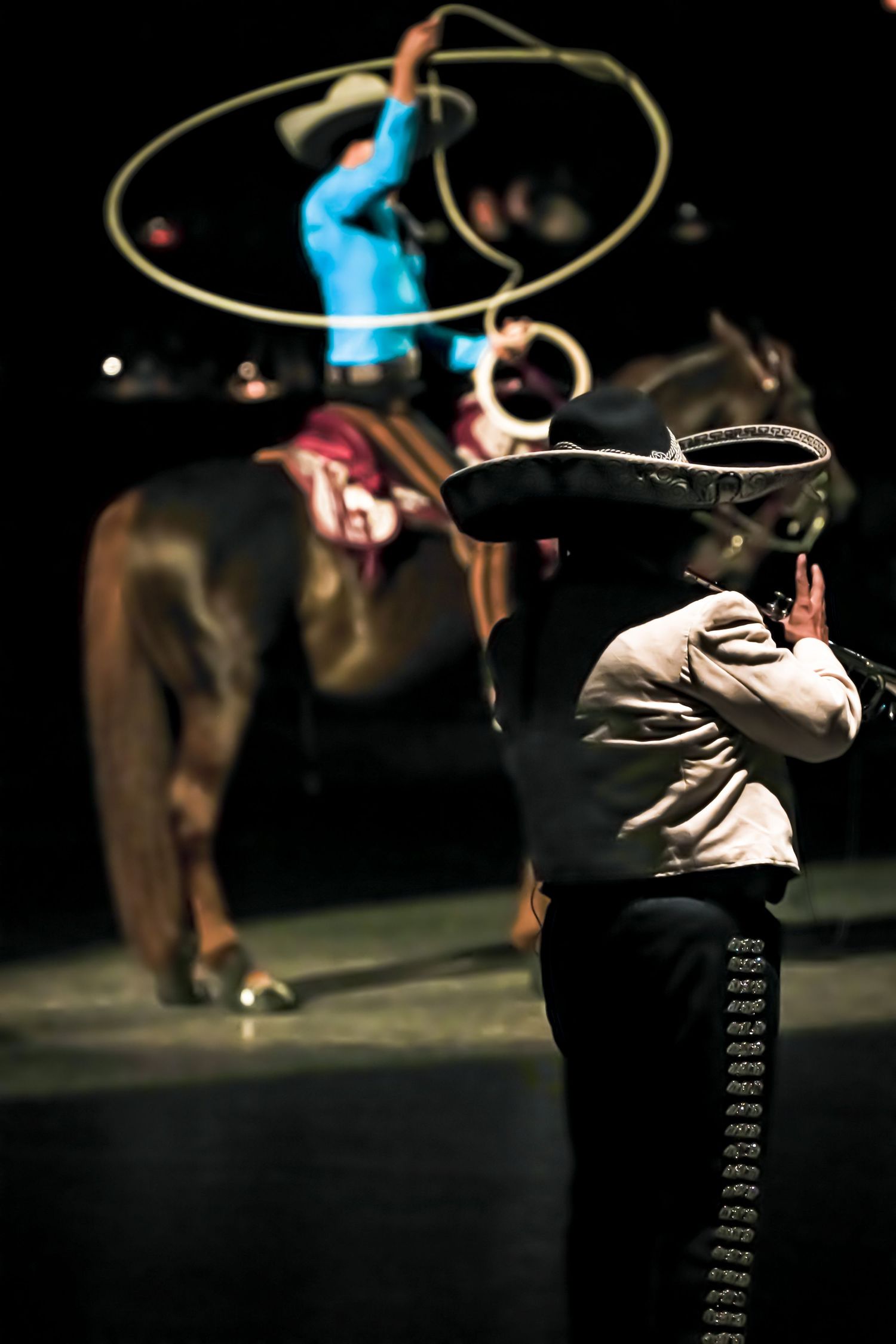 A charro in traditional attire watches a lasso performer on horseback during a Mexican rodeo show.