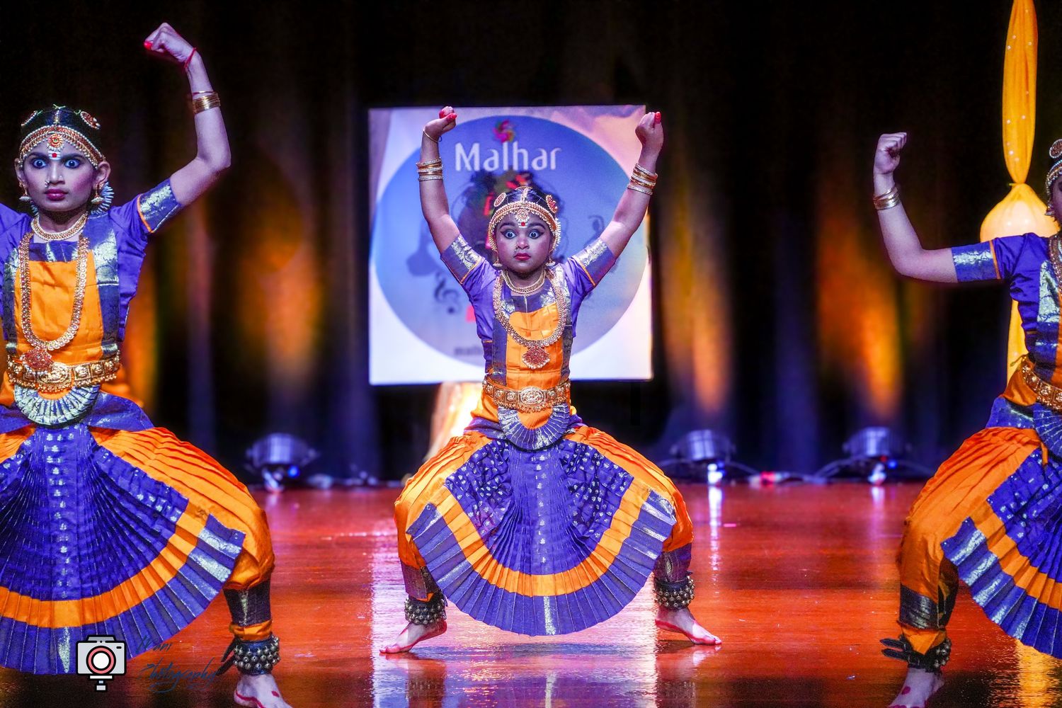 Three dancers perform Bharatanatyam in vibrant blue and orange costumes on stage at Malhar cultural festival.