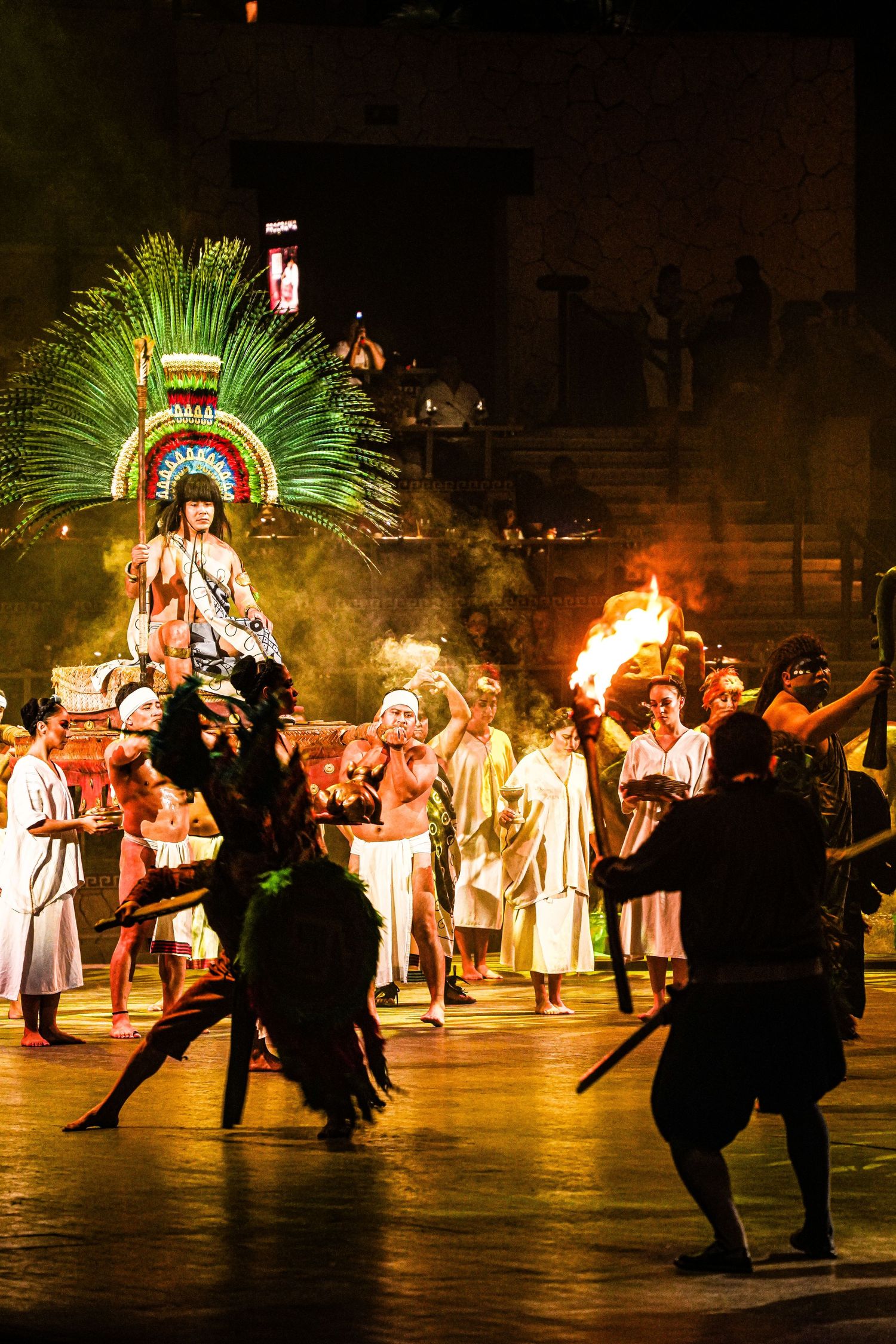 Aztec performers in traditional costumes with feathered headdress and fire torches during a nighttime cultural show.