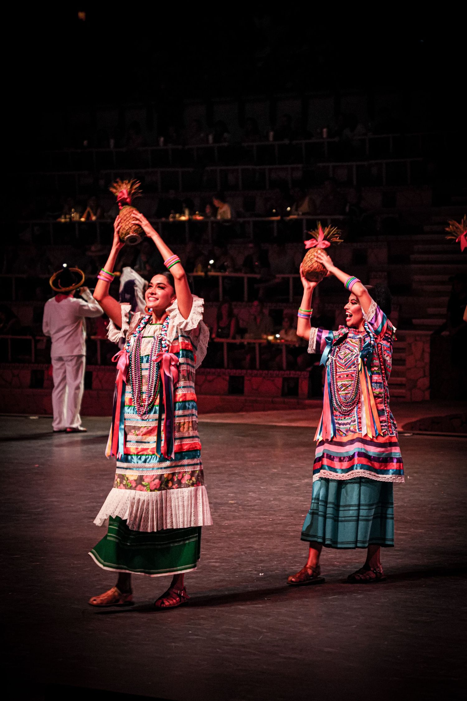 Two women in colorful traditional Mexican folk costumes perform a dance holding pineapples aloft on a stage.