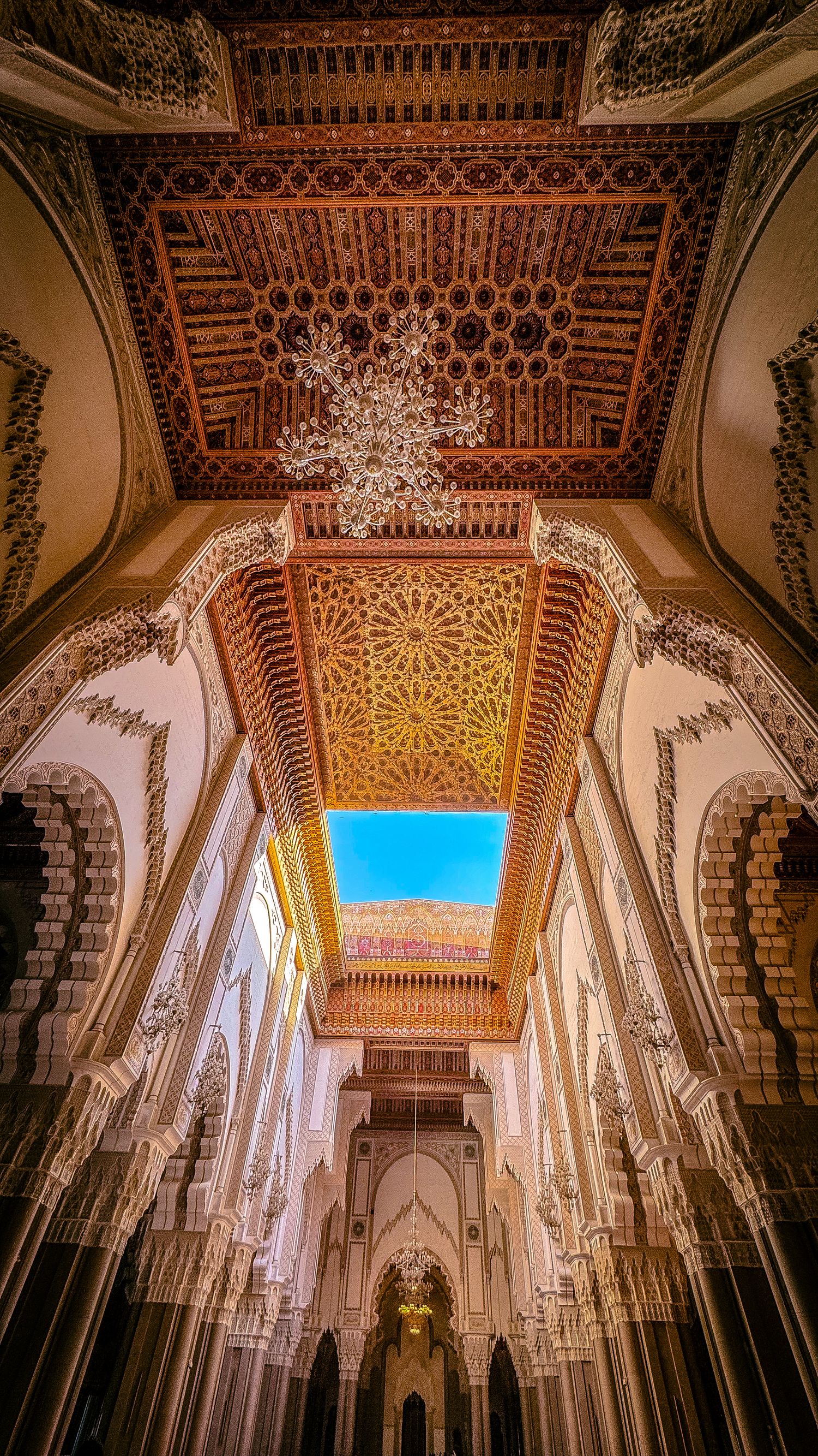 Ornate Moroccan mosque interior with intricate geometric ceiling, grand arches, chandelier, and open skylight revealing blue sky.