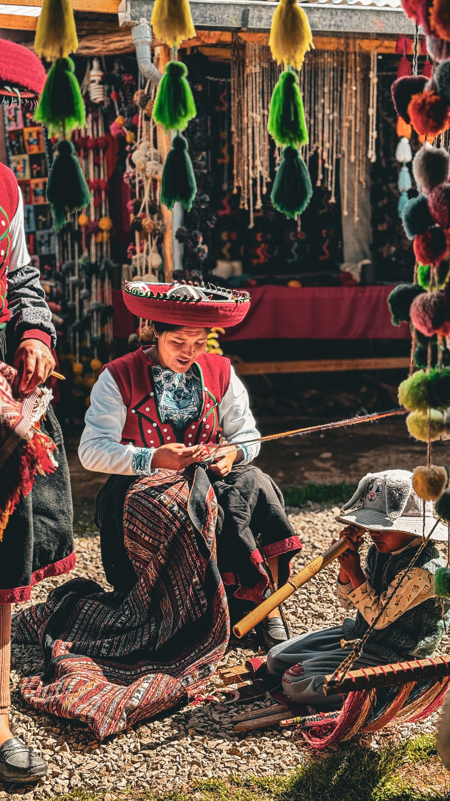 Two women in traditional Peruvian attire weave textiles at a colorful market stall with hanging tassels and crafts.