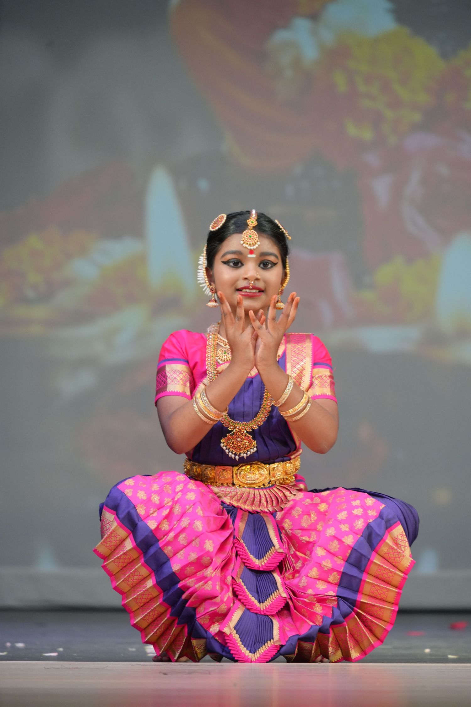 Young girl performing Bharatanatyam classical Indian dance in pink and purple silk saree with gold jewelry on stage.