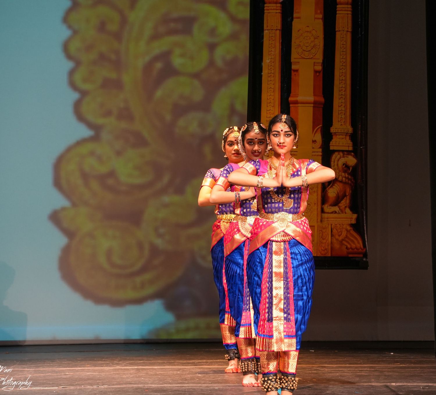 Three dancers perform Bharatanatyam in blue and pink silk costumes on a decorated stage.