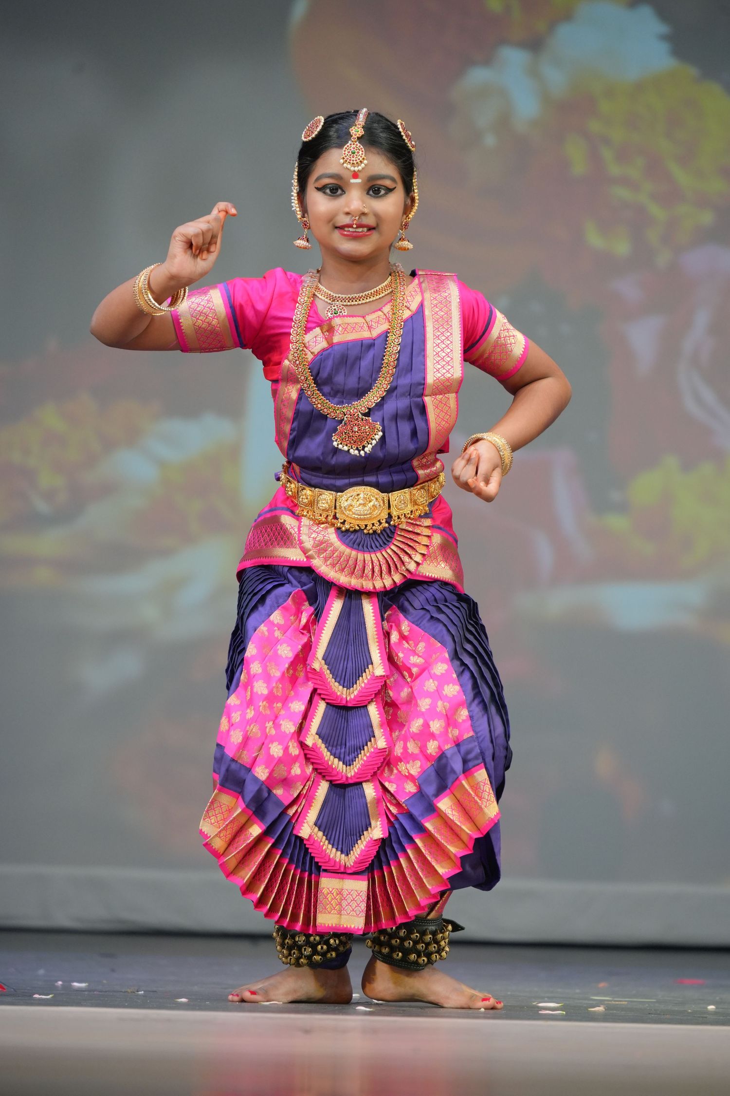 Young girl performing Bharatanatyam classical Indian dance in vibrant pink and purple silk costume with gold jewelry on stage.
