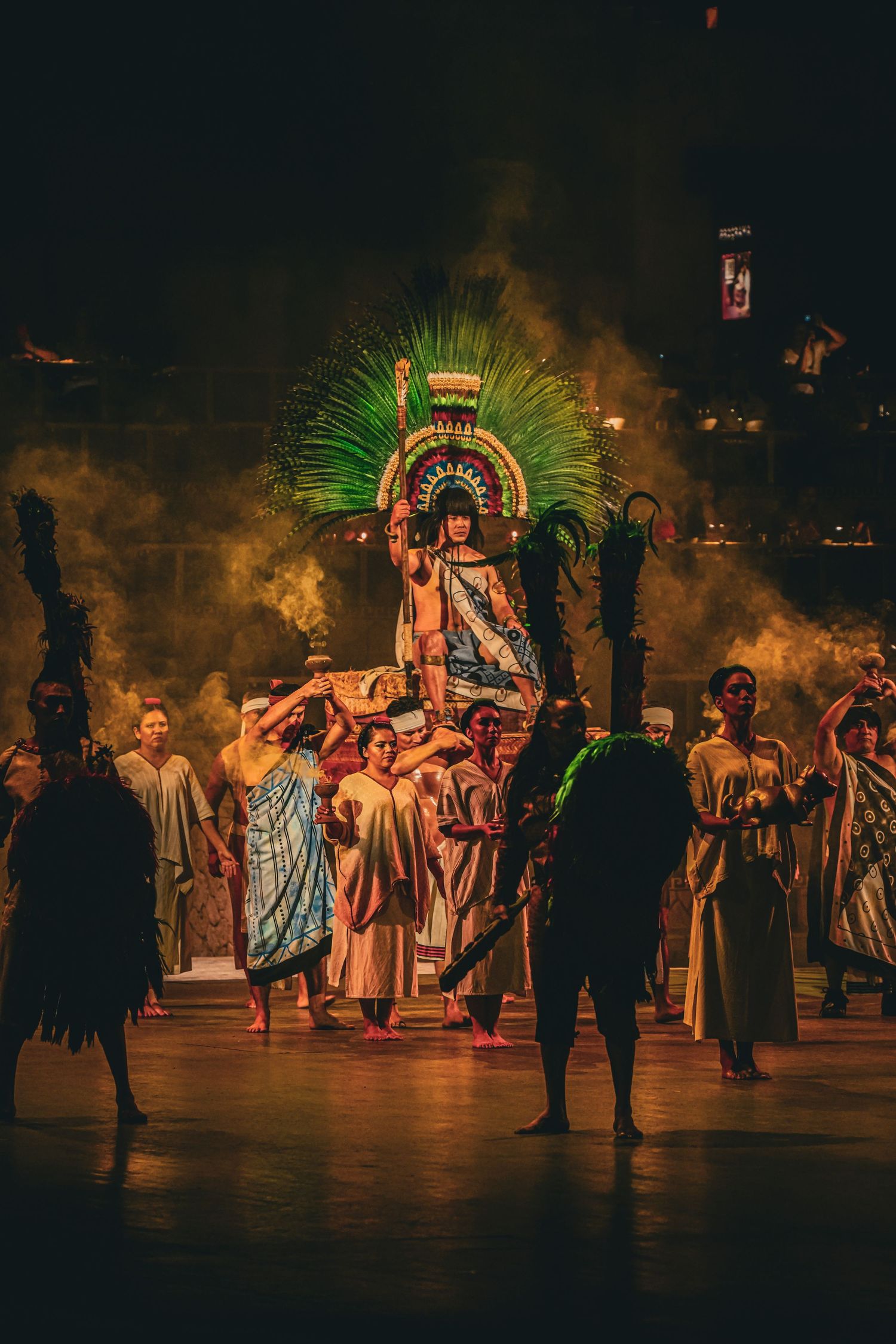 Aztec emperor carried on throne by performers in elaborate costumes with green feathered headdress during theatrical stage production.