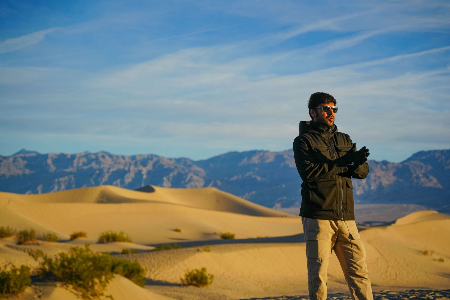 Man standing in vast desert sand dunes with mountain backdrop under blue sky, wearing jacket and sunglasses.
