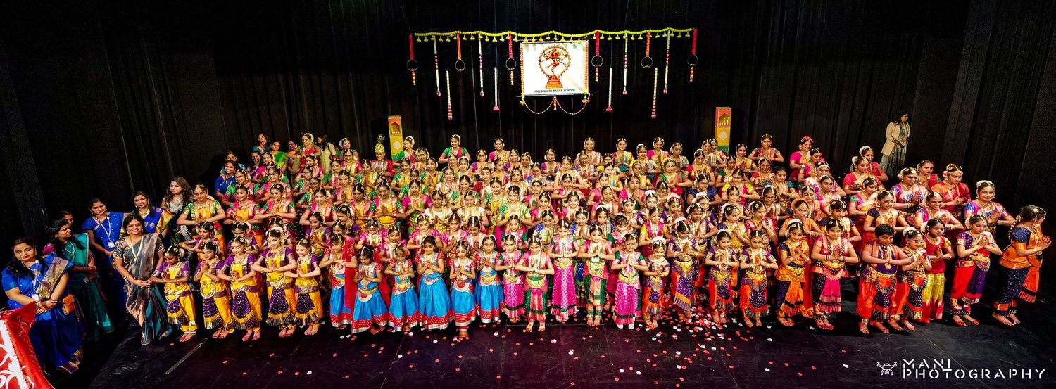 A large ensemble of Indian classical dancers in colorful traditional costumes posing on a decorated performance stage.