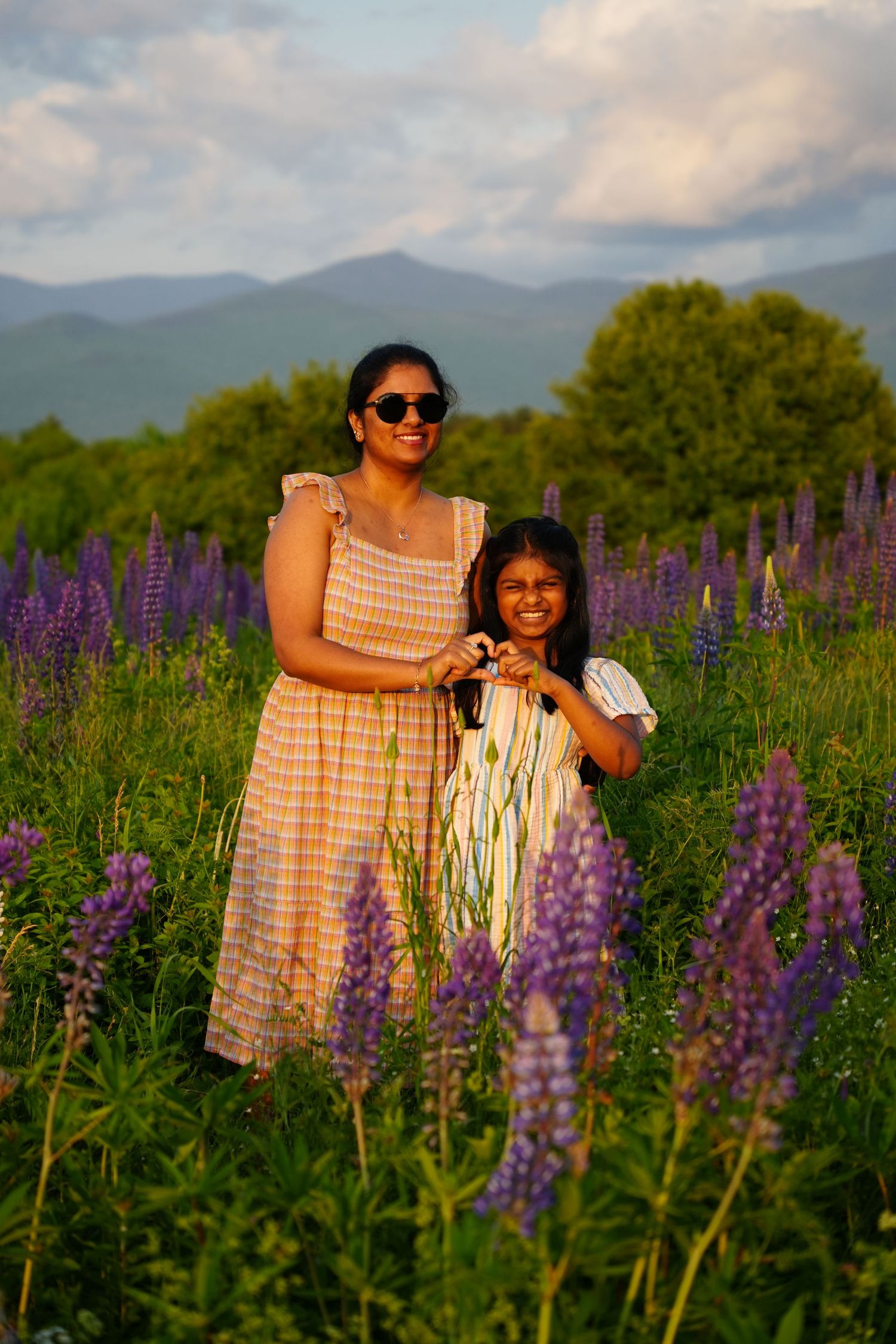 Woman and girl smile in purple lupine field with mountain backdrop at golden hour.