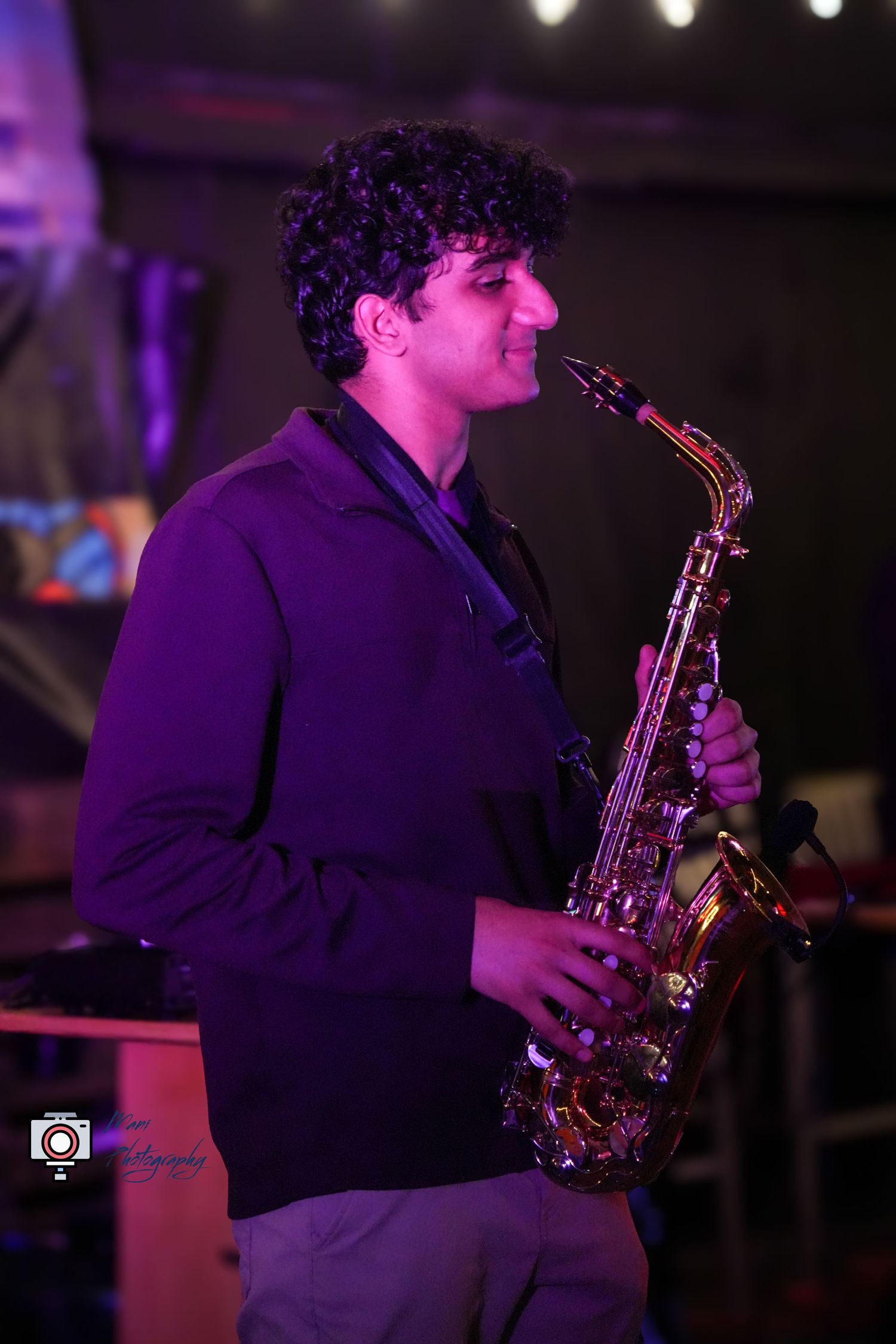 A young curly-haired musician plays a shiny alto saxophone under purple stage lighting at a live performance.