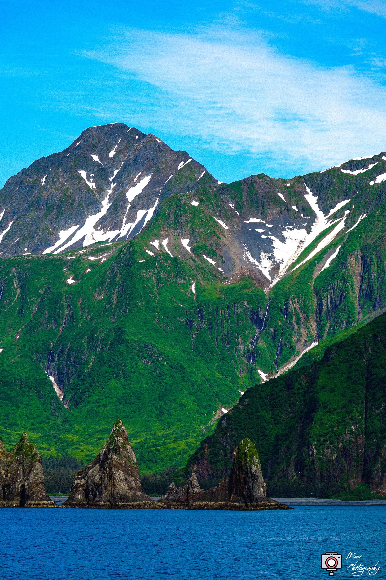 Stunning Alaskan mountain landscape with snow-capped peaks, green slopes, and blue ocean waters.