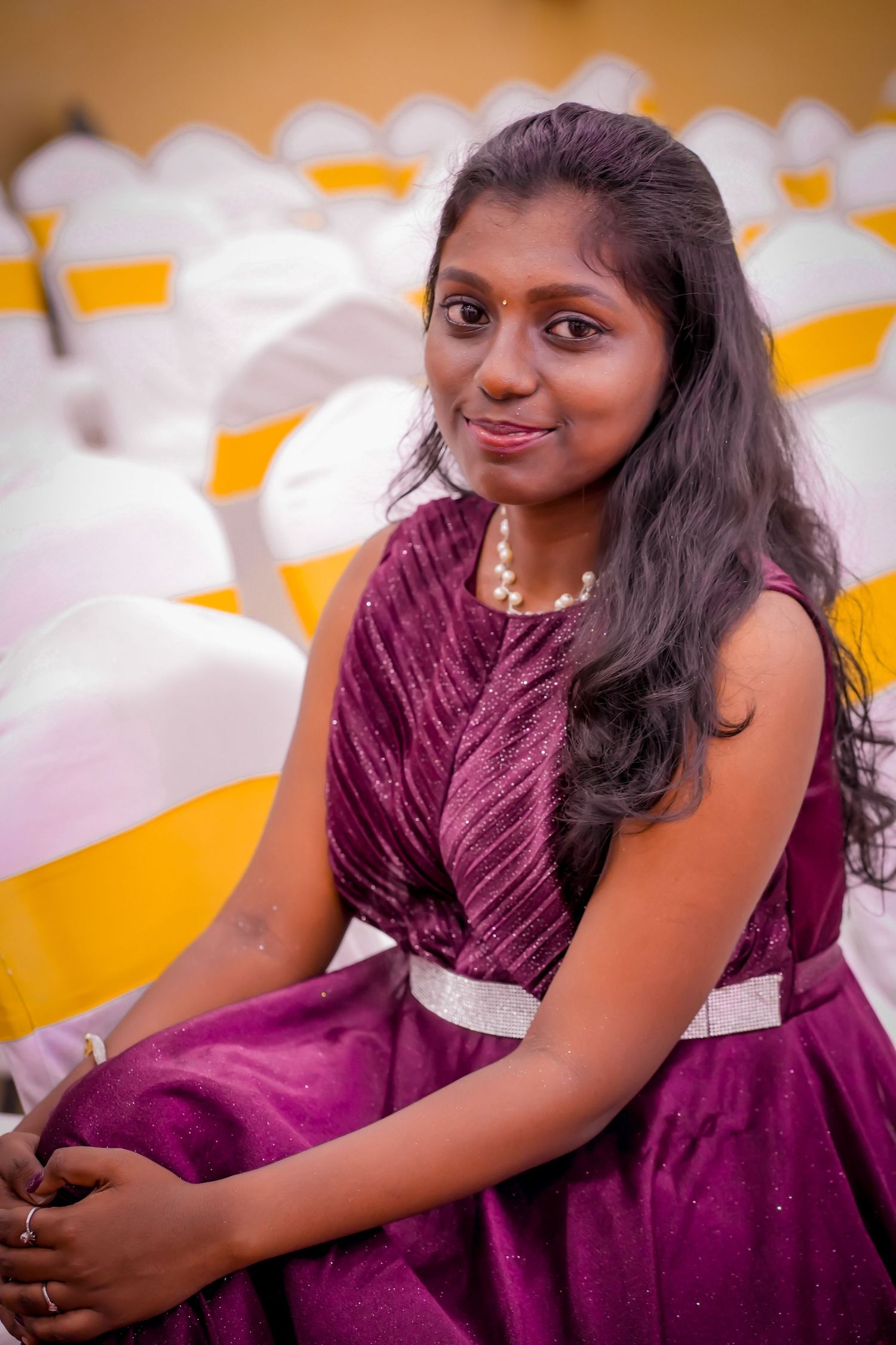 Young woman in purple dress with pearl necklace sitting among white chairs at an Indian event.