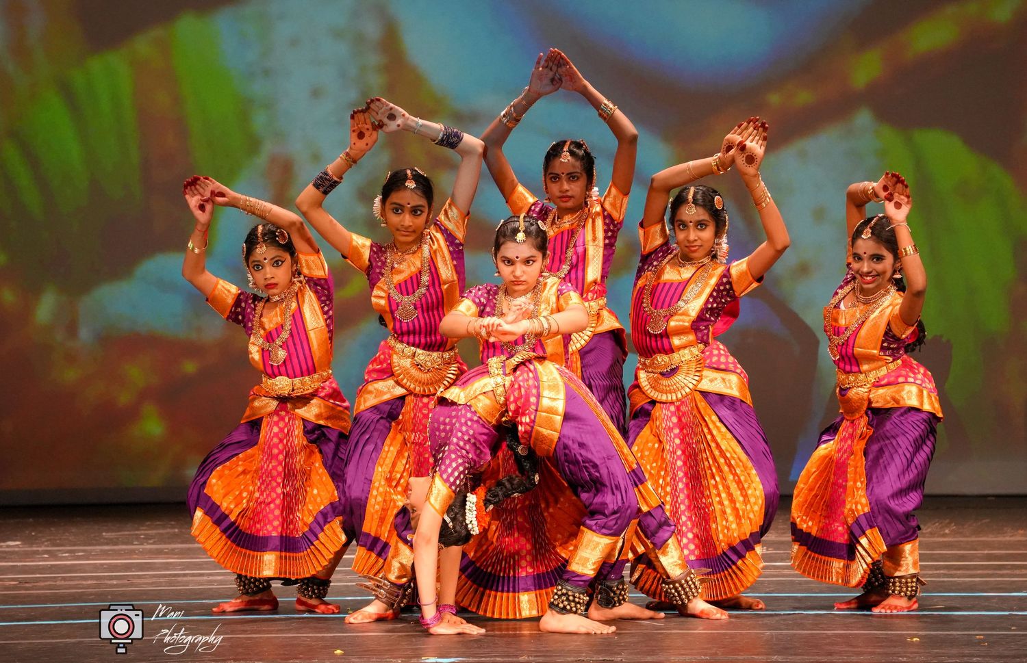 Indian classical dancers in vibrant orange and purple costumes perform Bharatanatyam on stage.