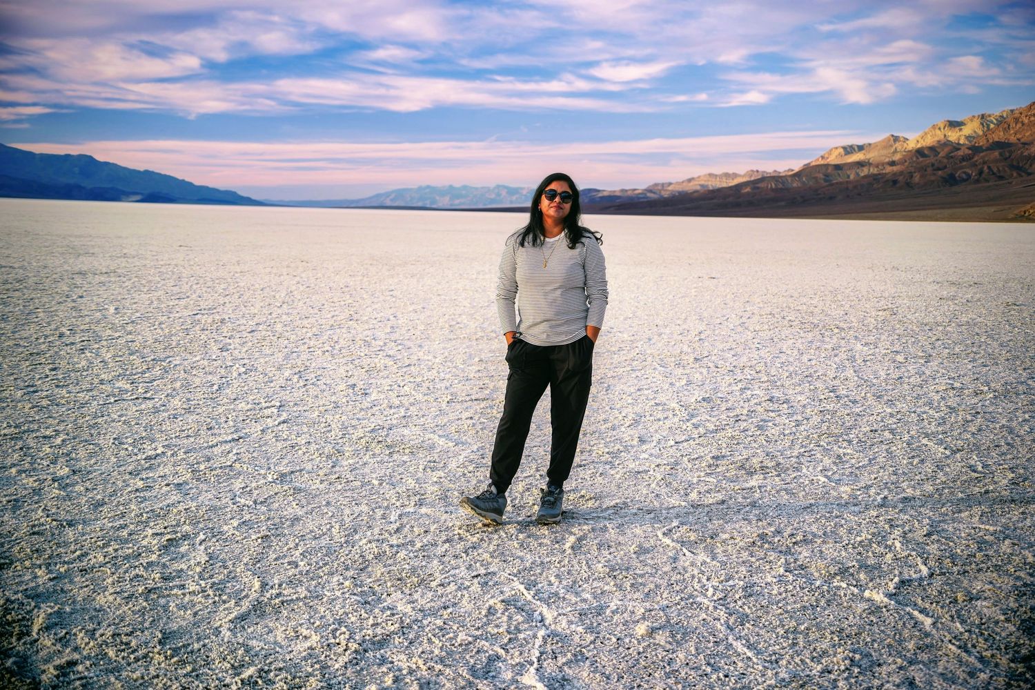 Person standing on vast white salt flats in Death Valley, wearing casual clothes and sunglasses, with mountains in background.