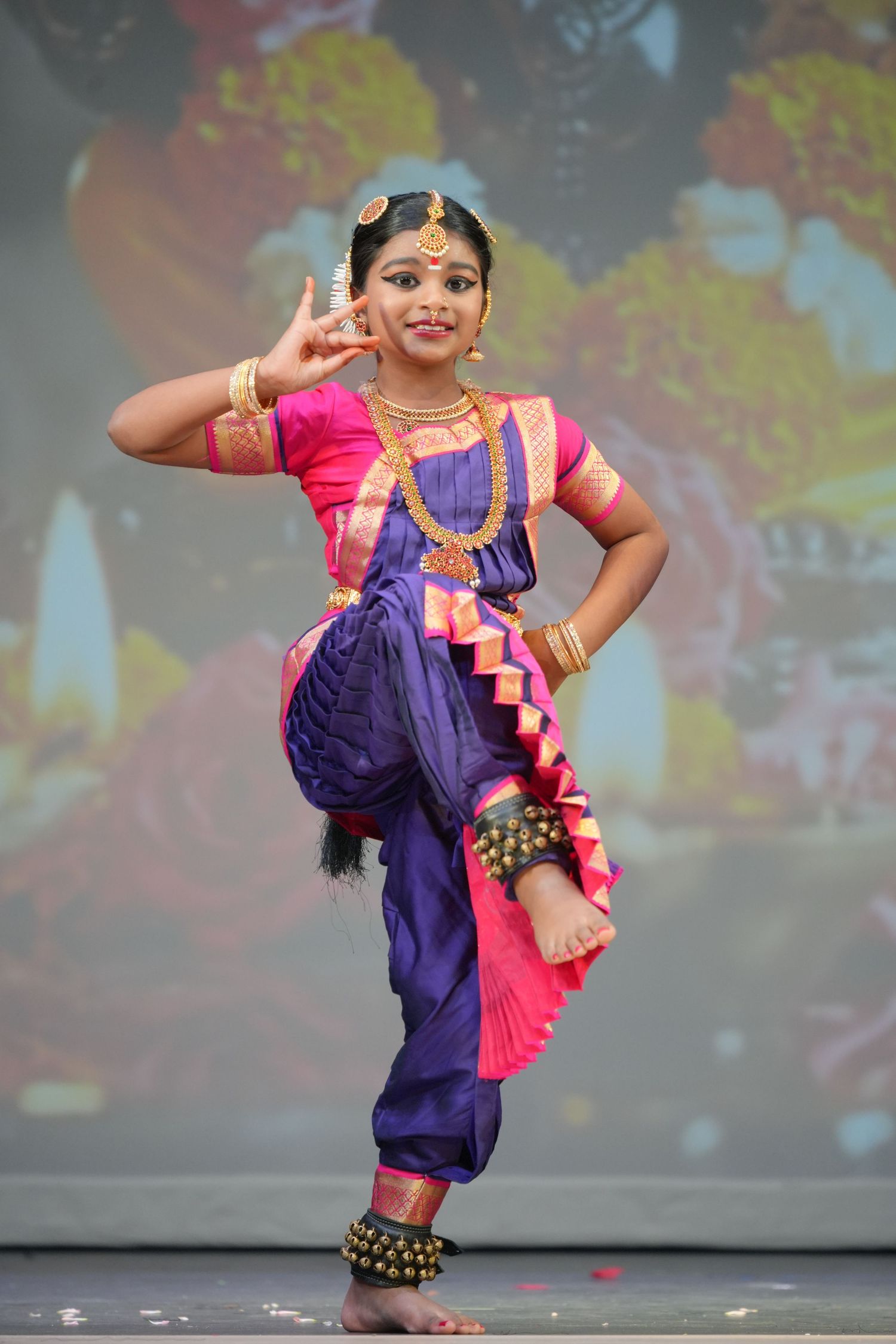 Young girl performing Bharatanatyam classical Indian dance in traditional purple and pink costume with gold jewelry.