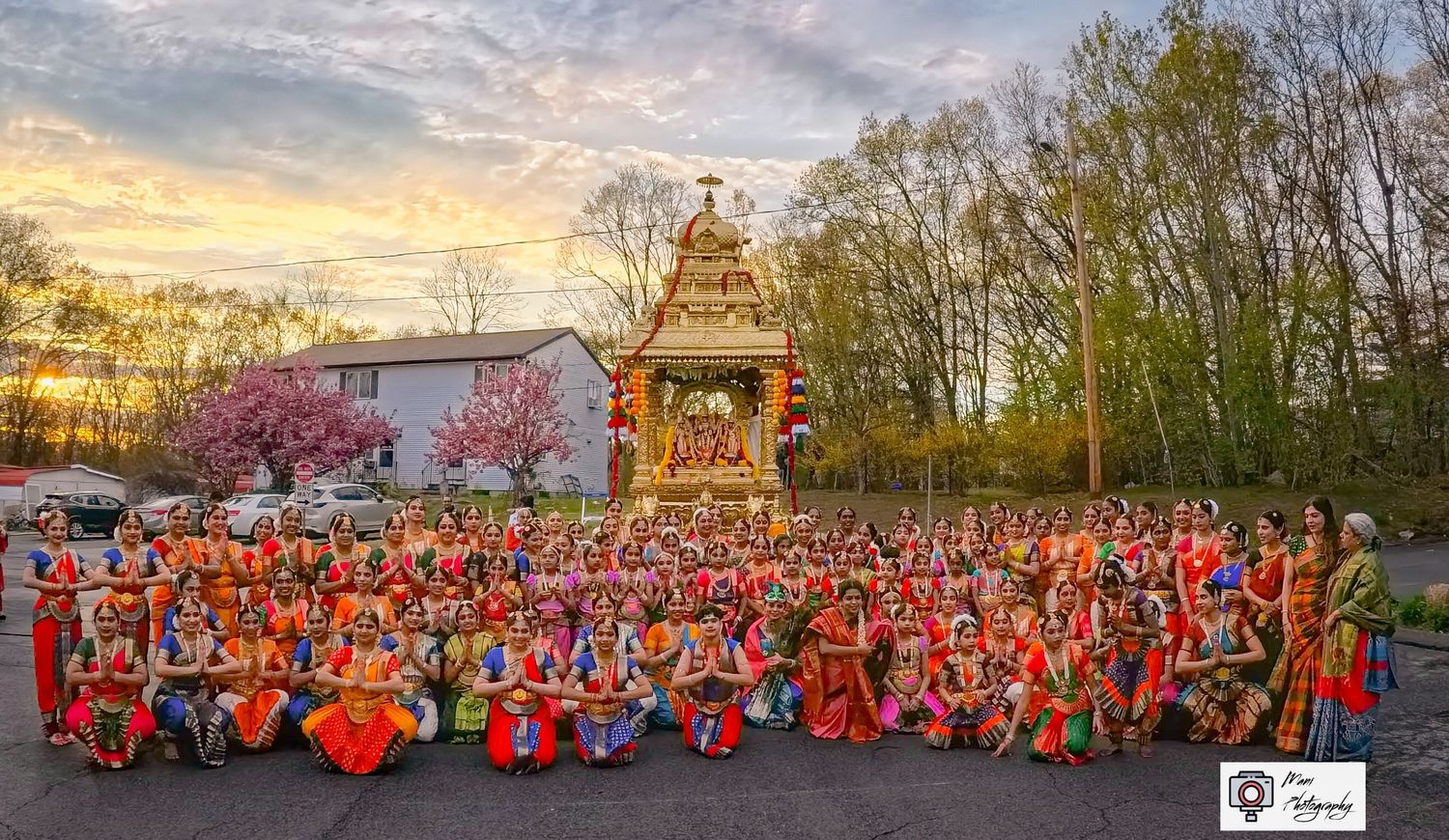 Large group of colorfully dressed dancers gathered before ornate Hindu temple at sunset.
