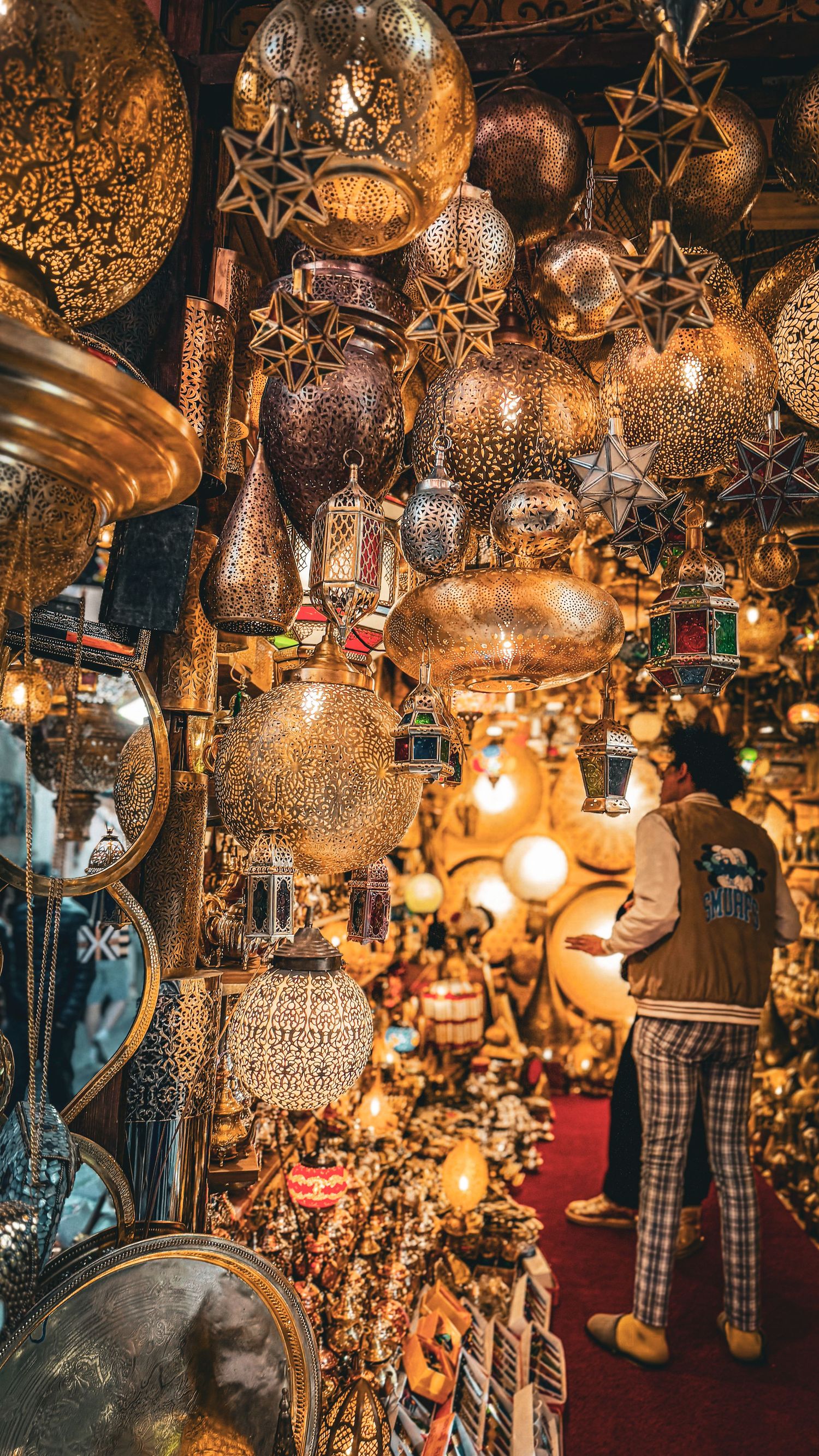 A person browses ornate golden Moroccan lanterns and star-shaped lamps hanging in a vibrant souk market stall.