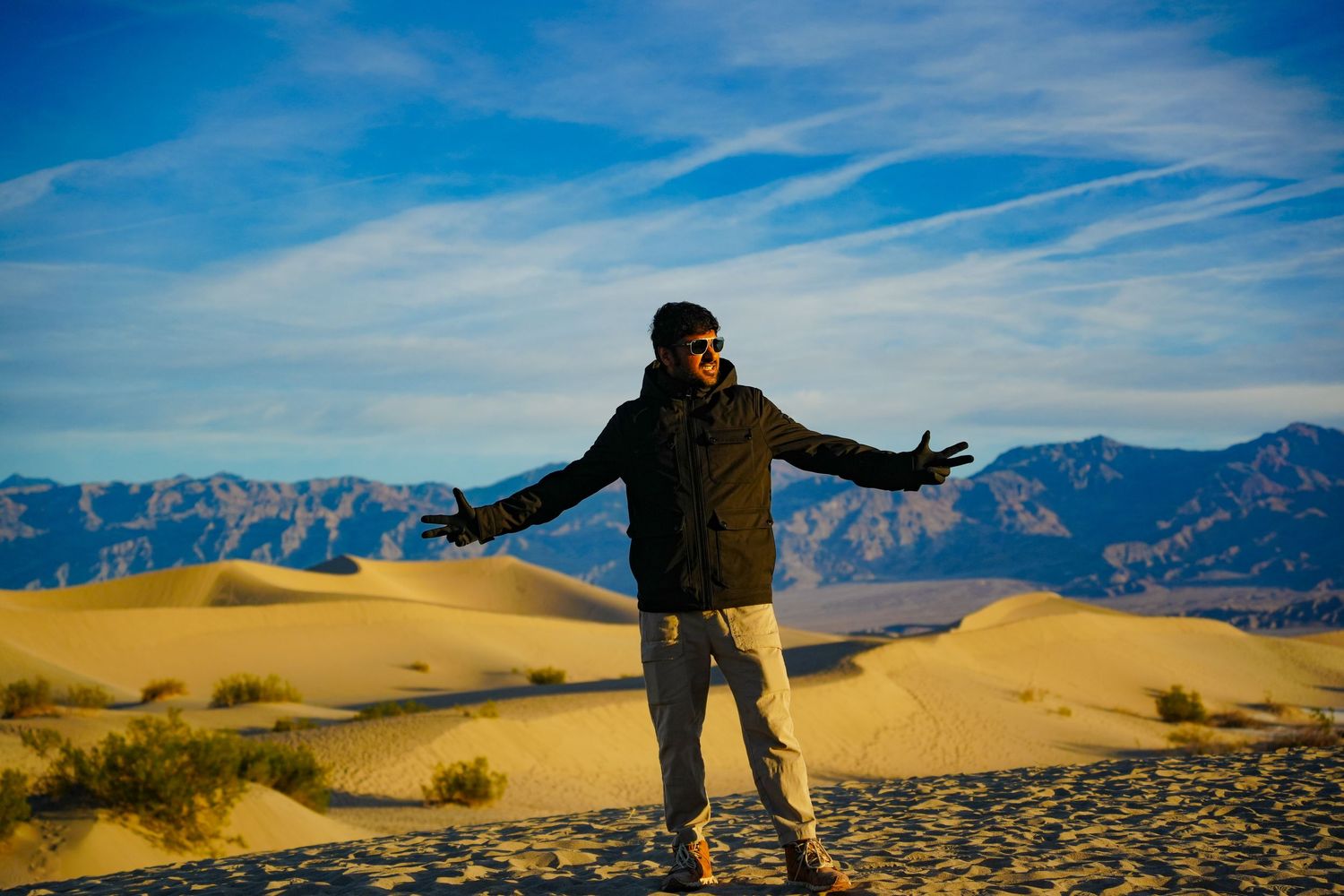 Man with arms outstretched standing on golden sand dunes with mountain backdrop and blue sky in desert landscape.