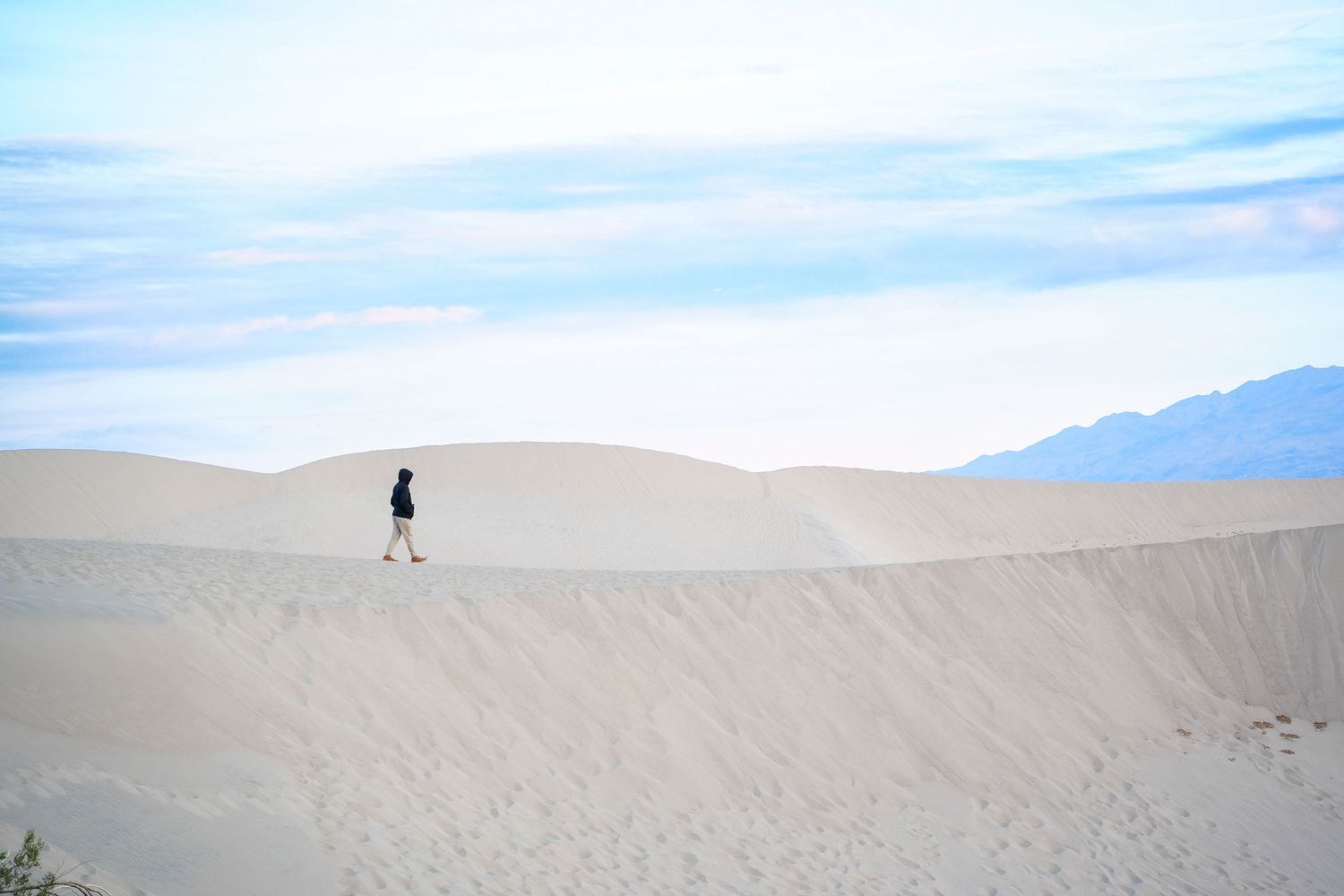 A lone hiker walks along white sand dunes under a blue sky with mountains in the background at Death Valley.
