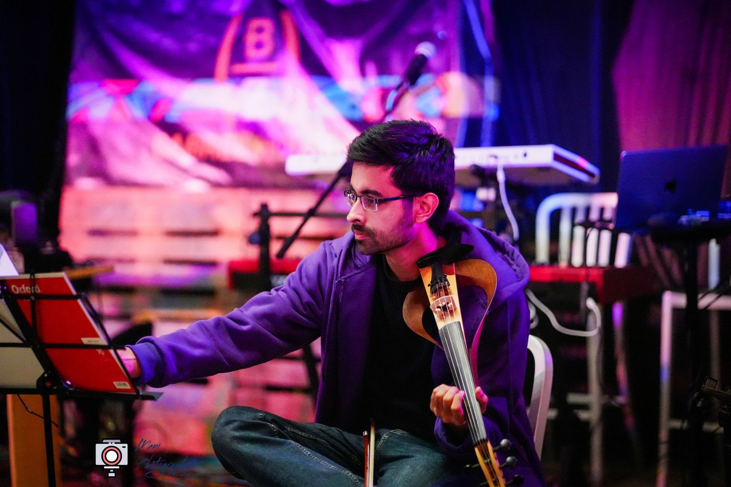 Musician playing cello on stage in purple jacket at colorful live music event.
