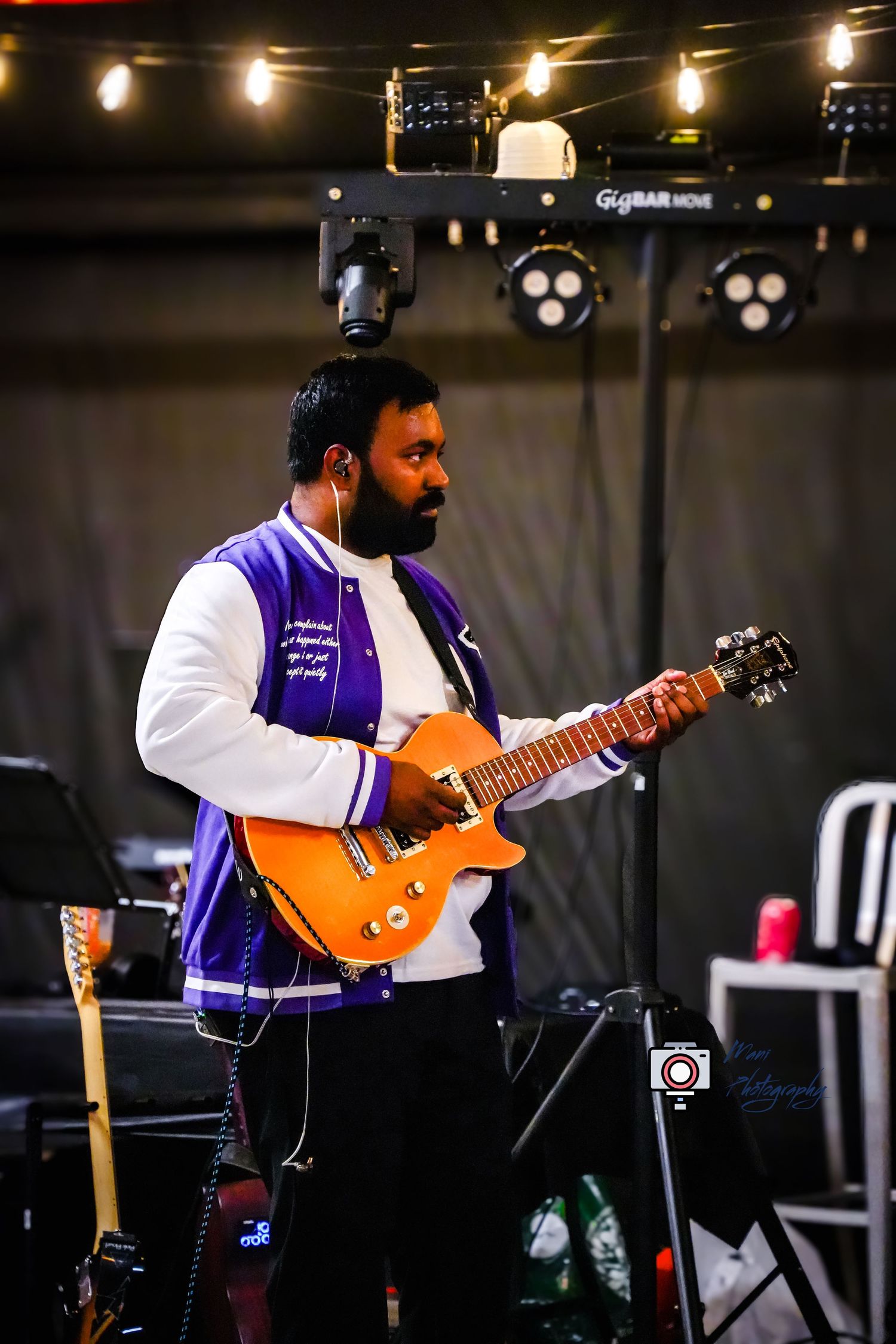 Man in purple varsity jacket playing orange electric guitar on stage with colorful lights at live music event.