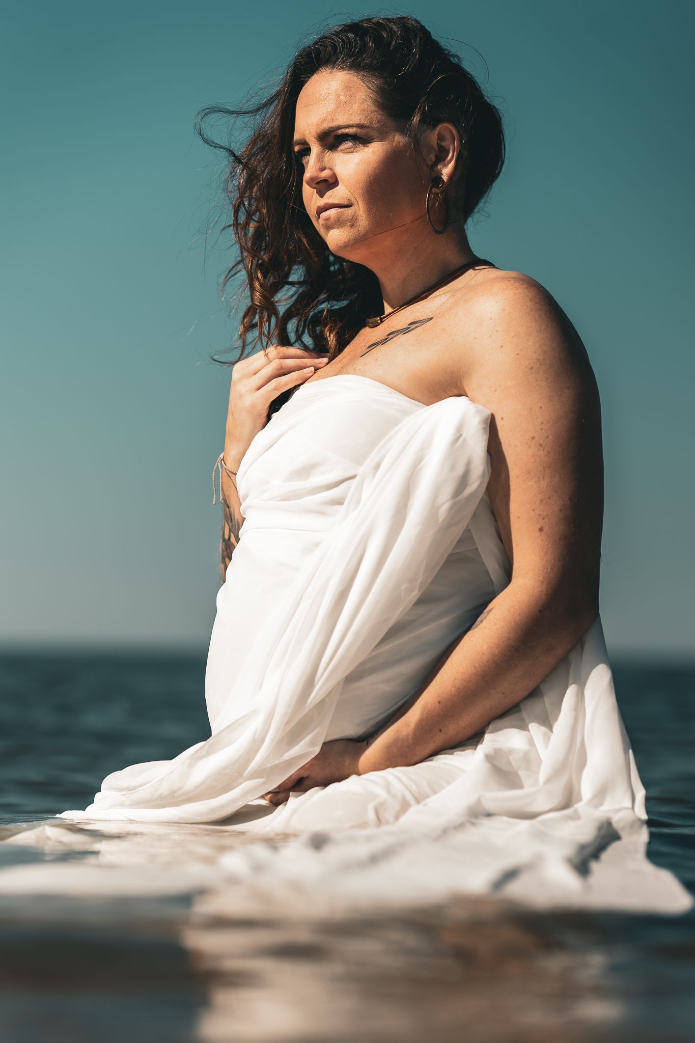 Pregnant woman wrapped in white fabric sitting in ocean water gazing into distance on sunny day.