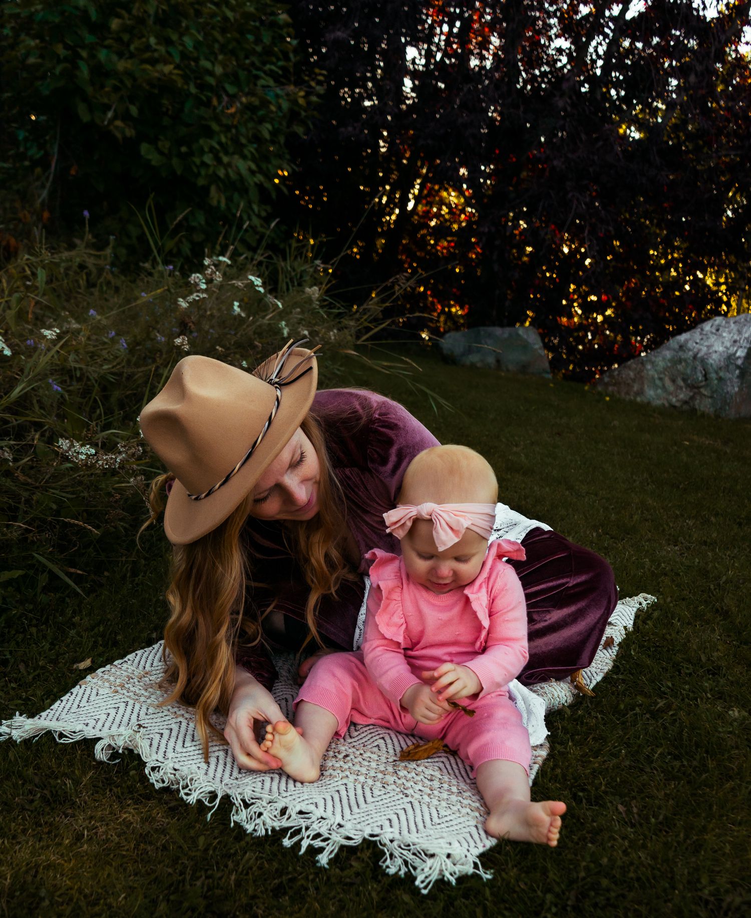 Mother in tan hat and burgundy dress sits on blanket outdoors with baby in pink outfit.