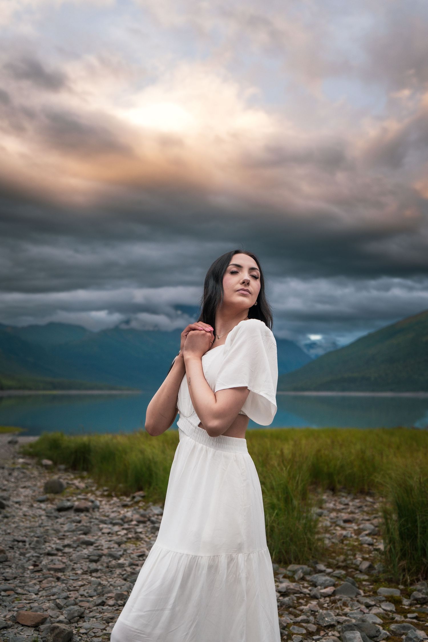 Woman in white dress poses by mountain lake under dramatic stormy sky at sunset.