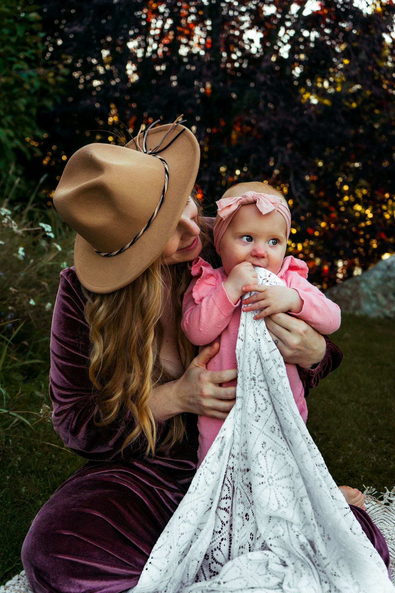 Blonde woman in tan hat and purple velvet dress holds baby girl in pink outdoors.