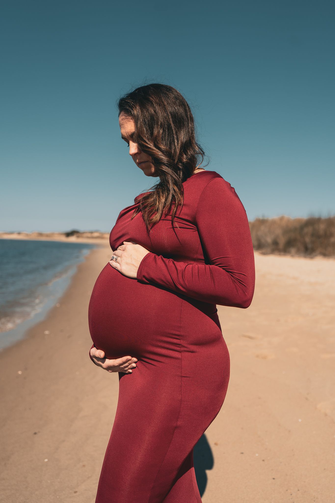 A pregnant woman in a burgundy red dress gently holds her baby bump while standing on a beach.