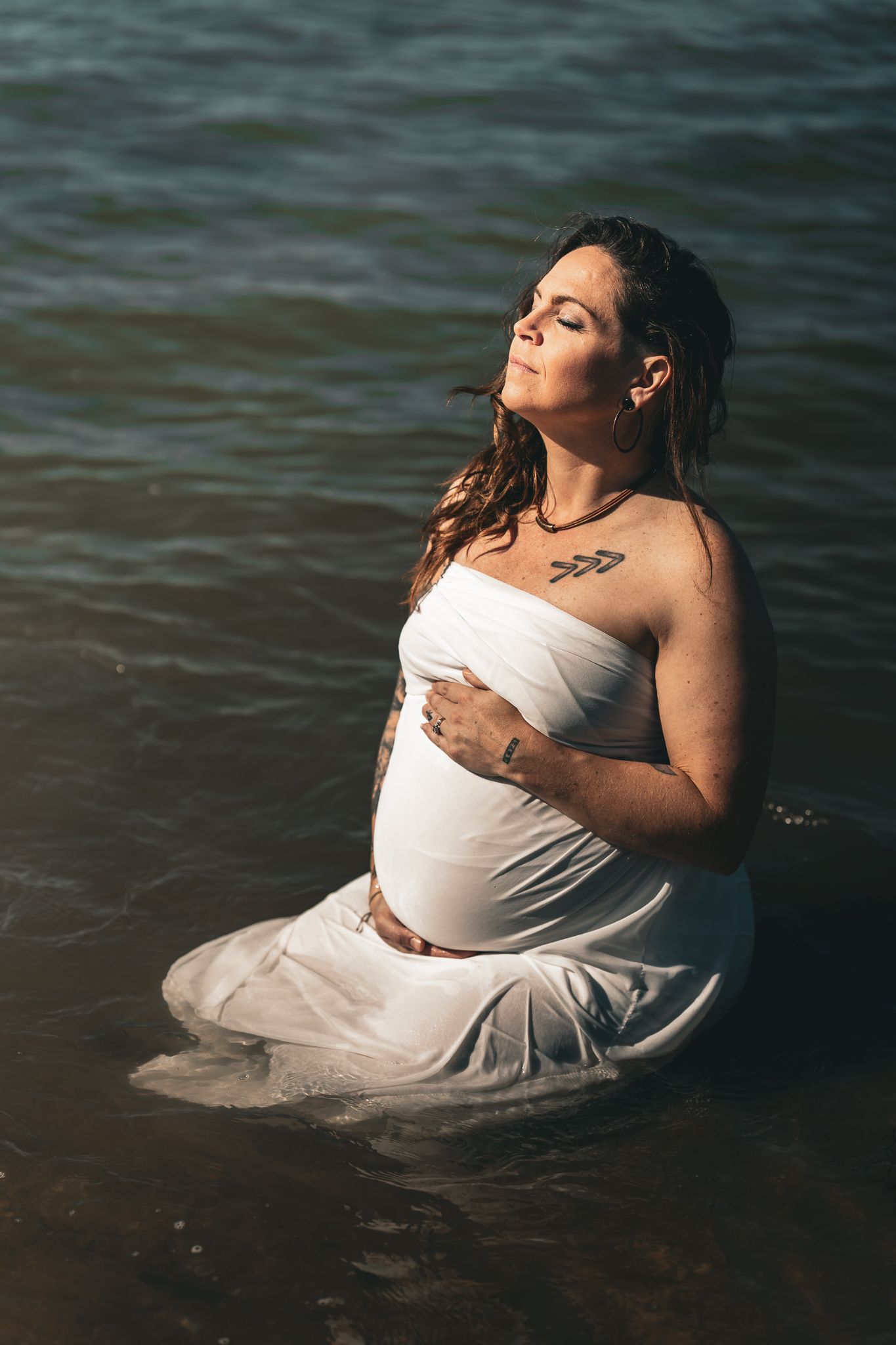 A pregnant woman in white fabric sits peacefully in ocean water with eyes closed in sunlight.