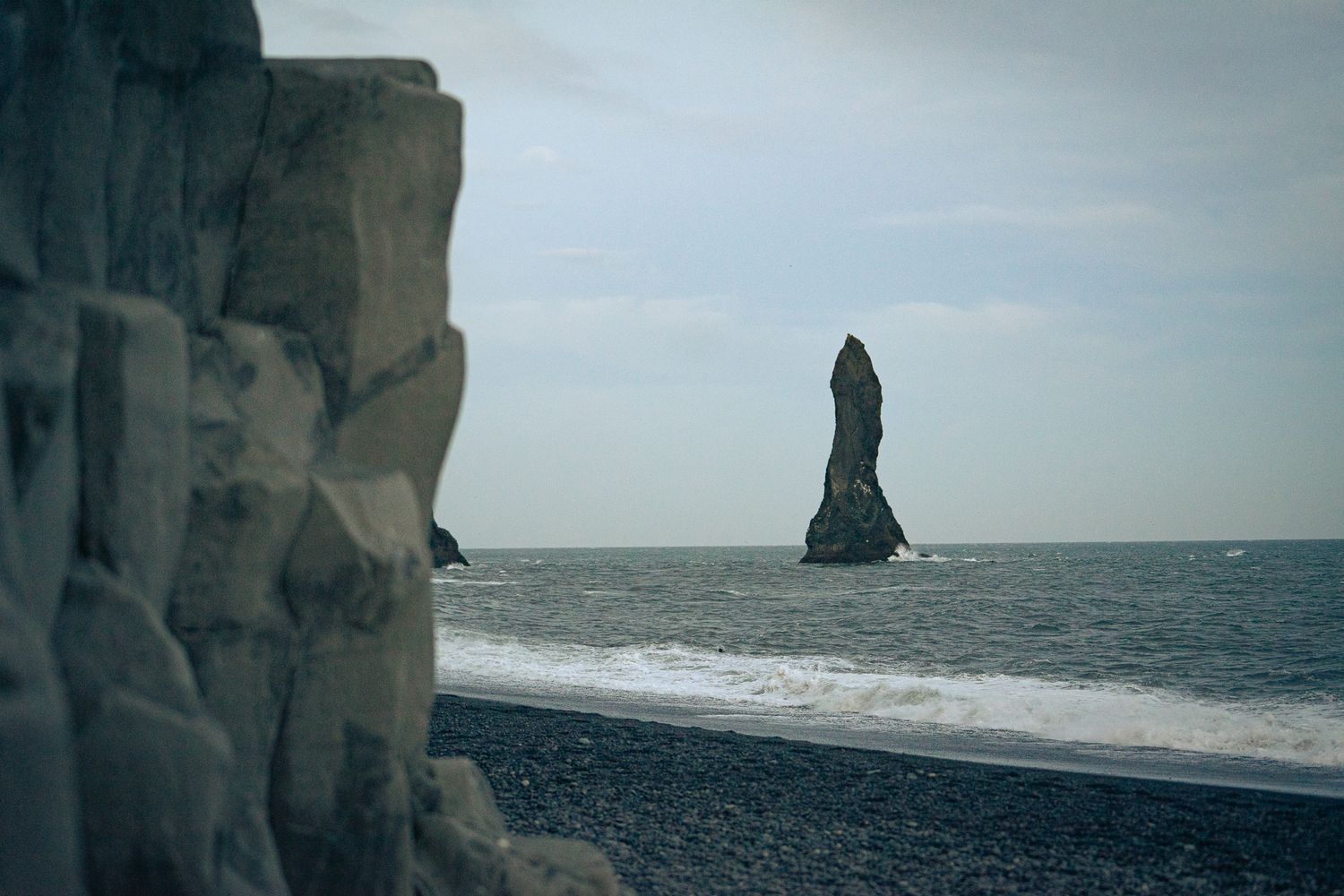 Black volcanic rock formations and sea stack rising from dark ocean waves on Iceland's black sand beach.